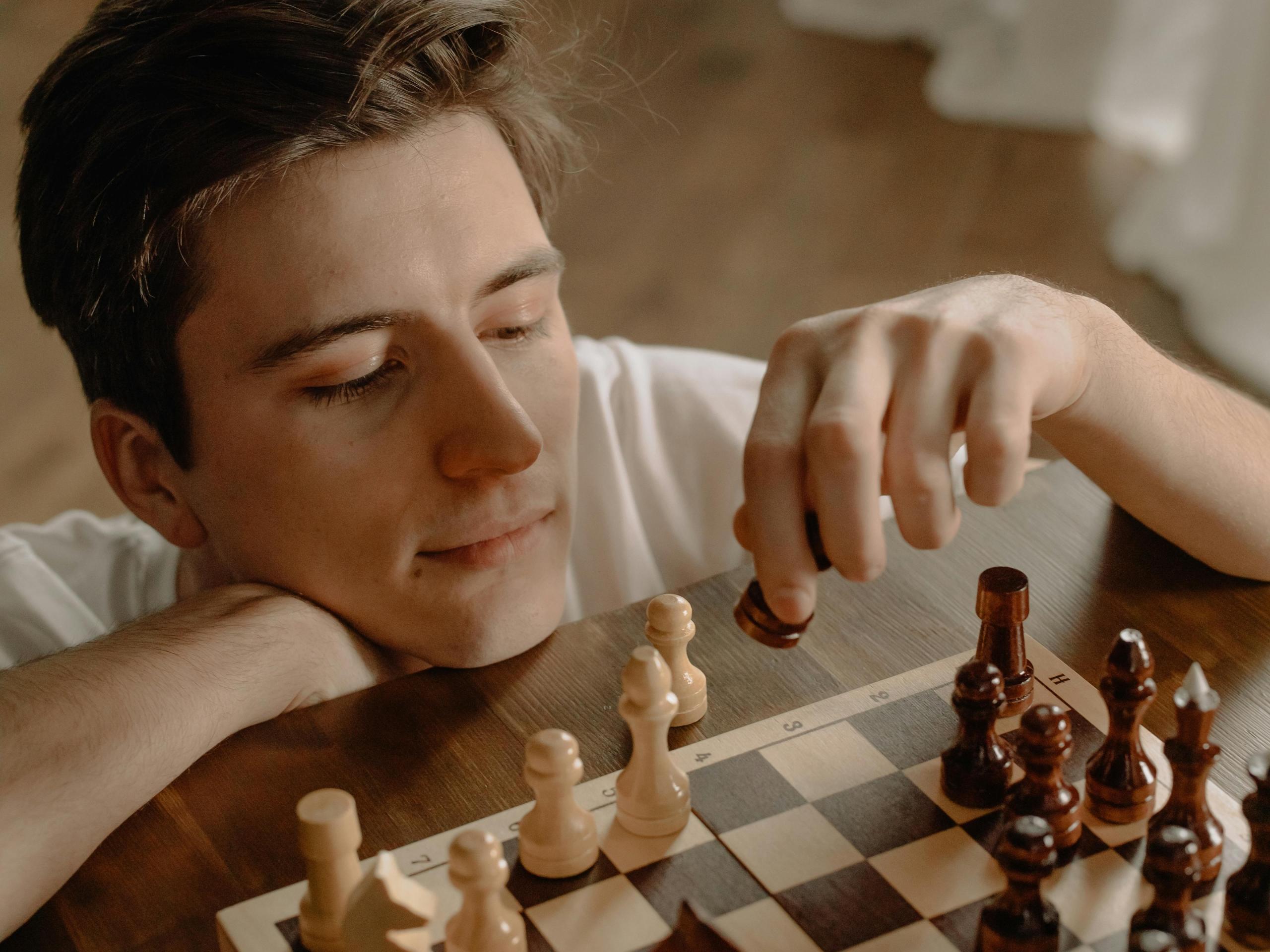 a man rests his chin on a table. He moves a chess piece on a board that is covered with dark and light chess pieces. 