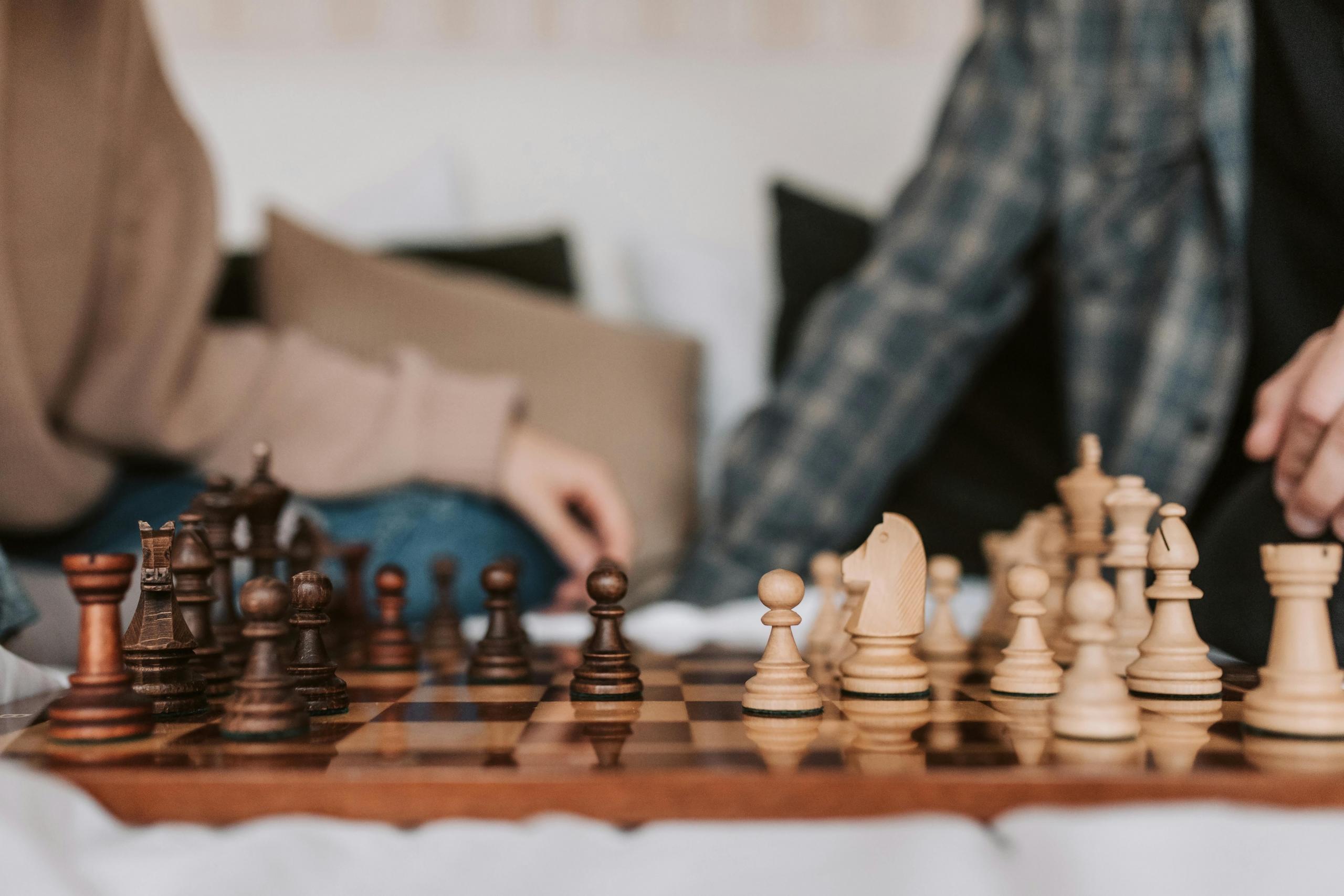 a wooden chess board is set up. In the background, two people sit on a couch. 
