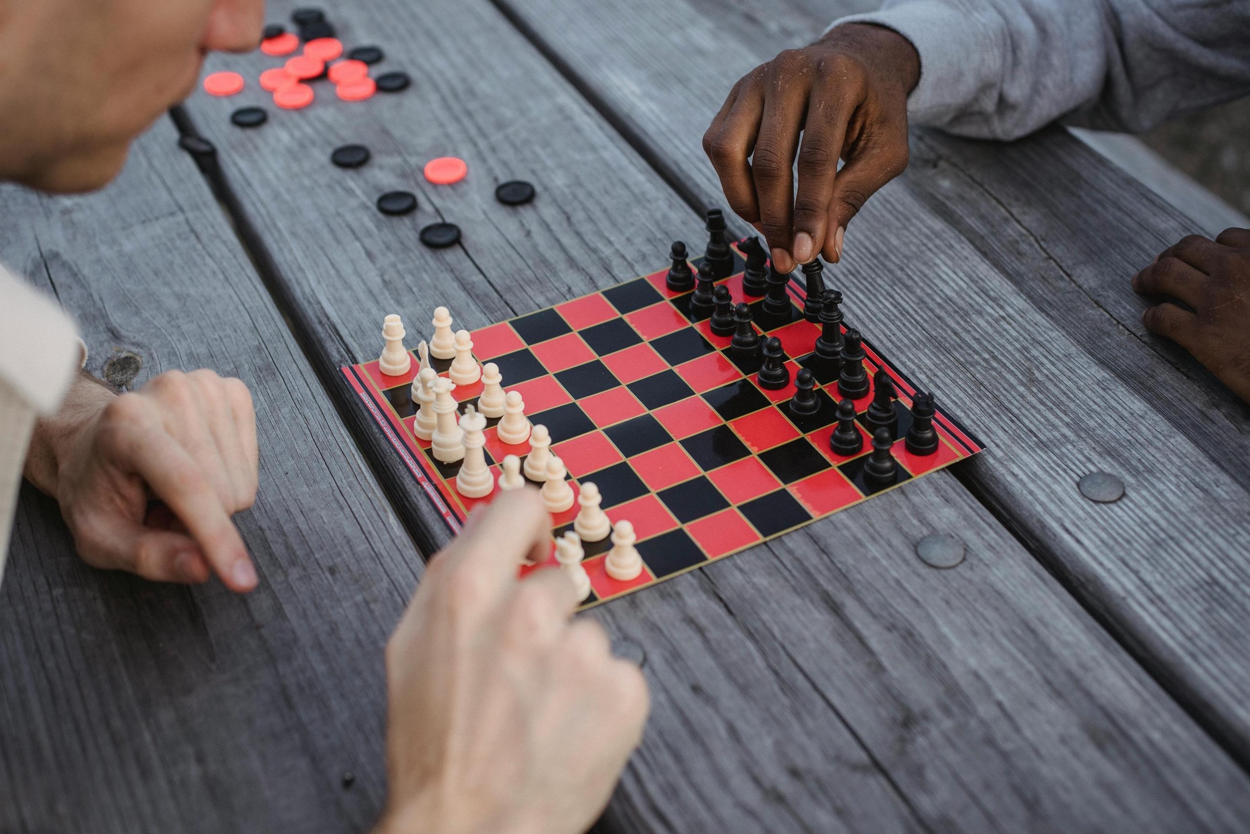 Two men play chess on a small board on a wooden picnic table. Checkers pieces are scattered on the table.