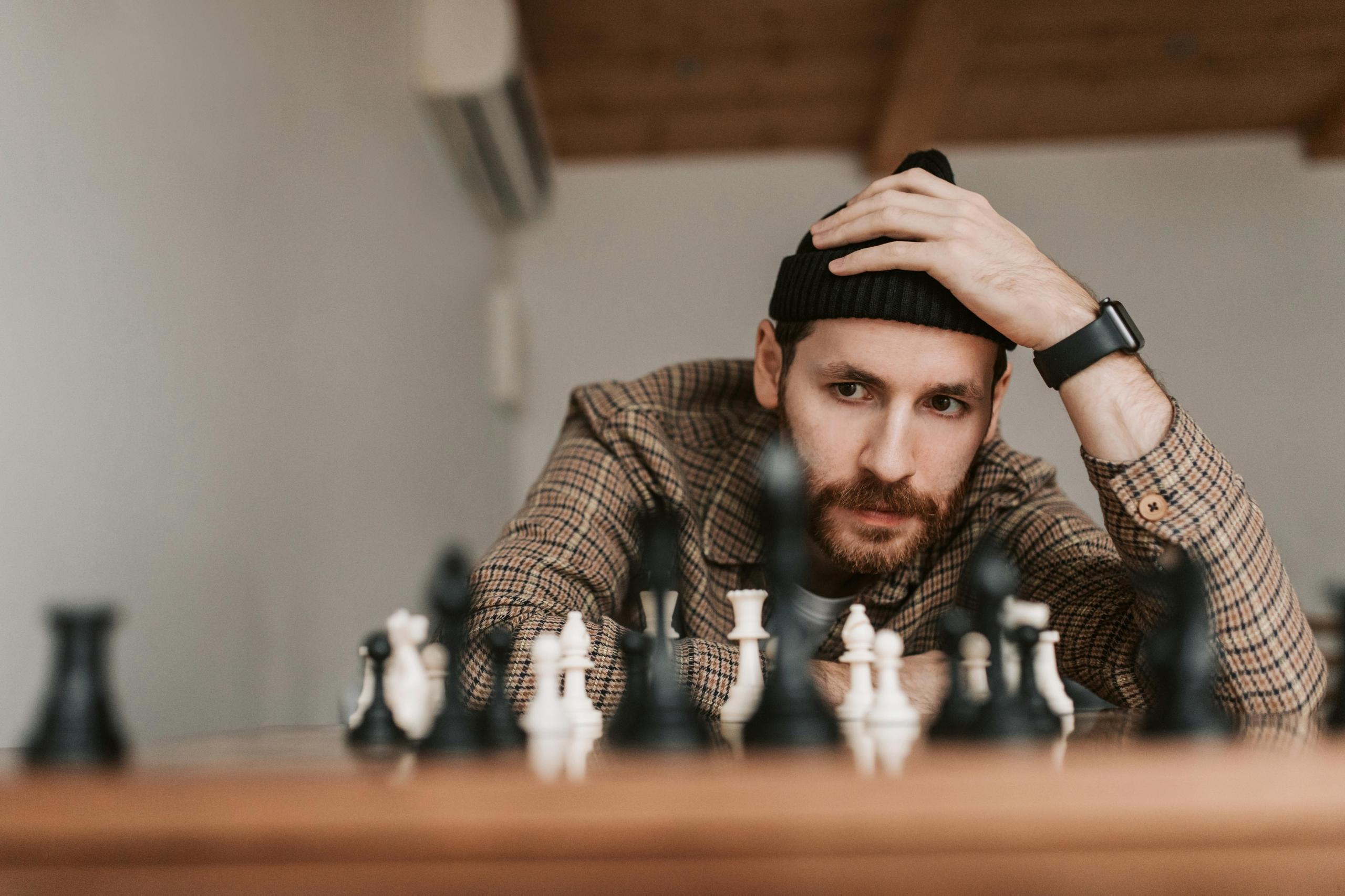 A man wearing a tuque looks at a chess board. His hand is holding his head.