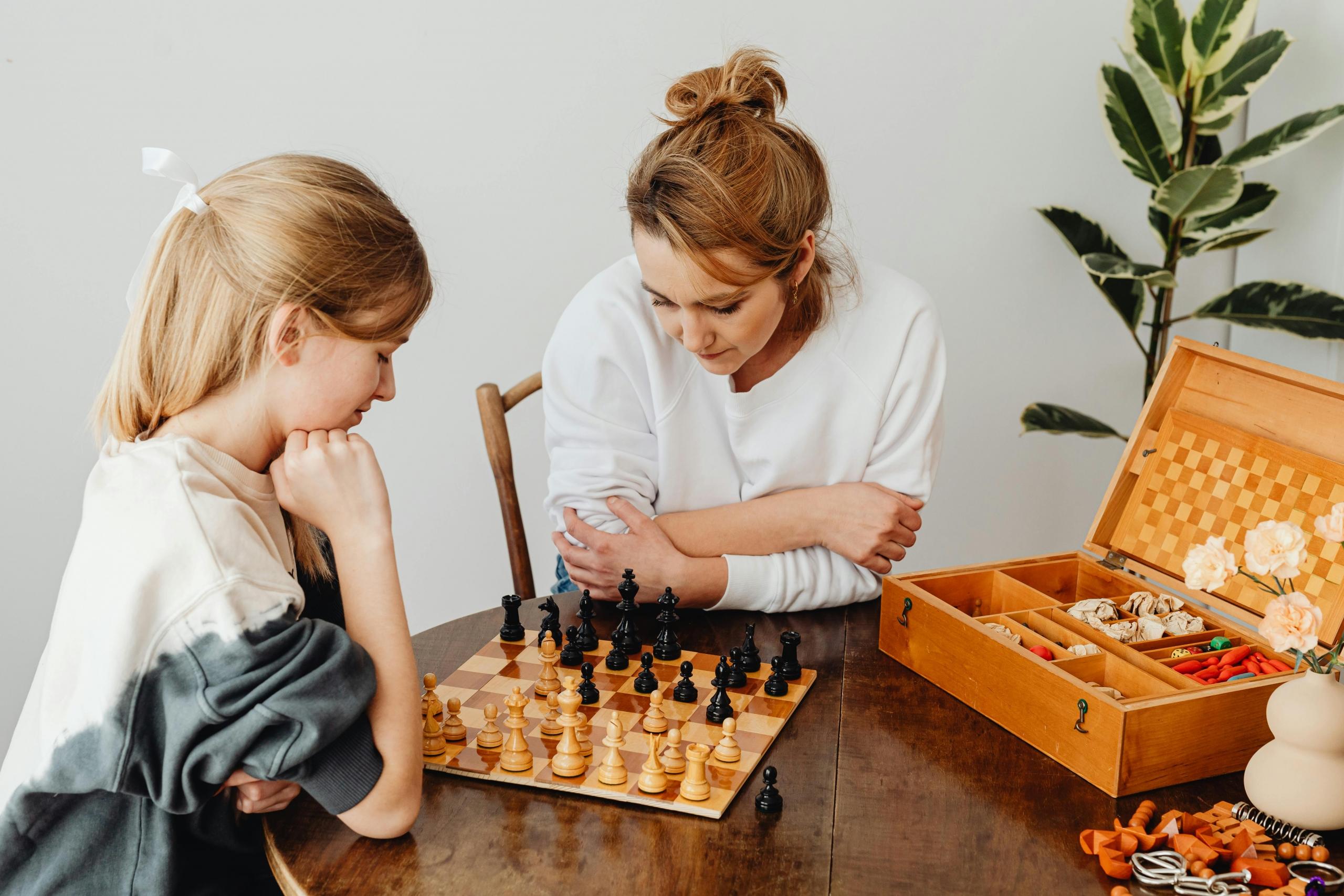 two women with light hair play chess on a table. There is a plant in the back and a wooden box that held the chess pieces and board.