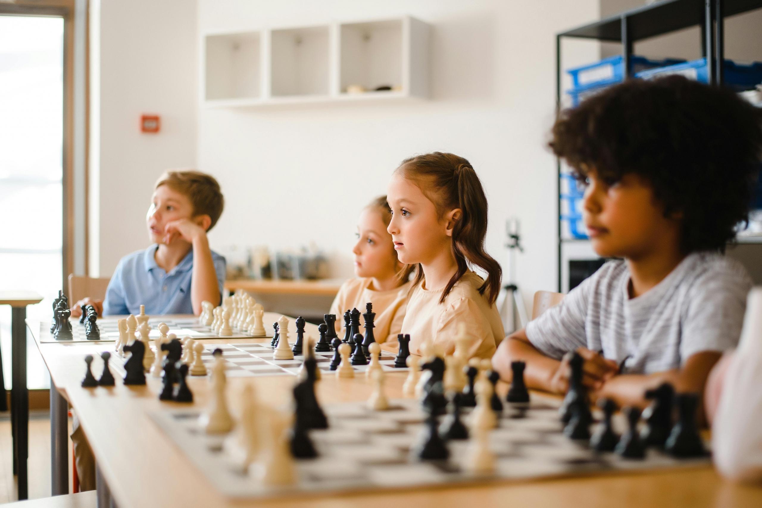 a group of children sit around chess boards, awating instruction.