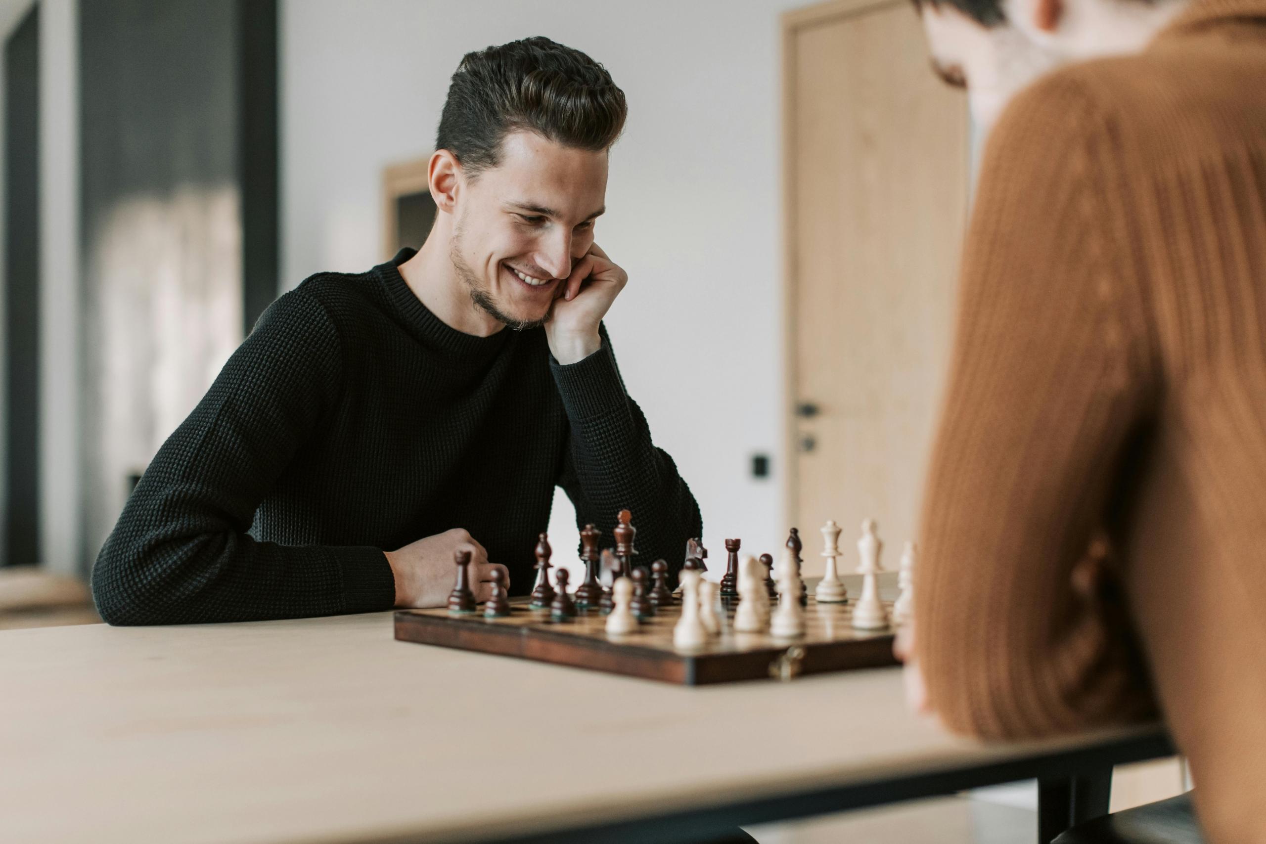 a man wearing a black shirt looks down at a chess board, smiling. An opponent in a brown sweater can be seen sitting across the table.