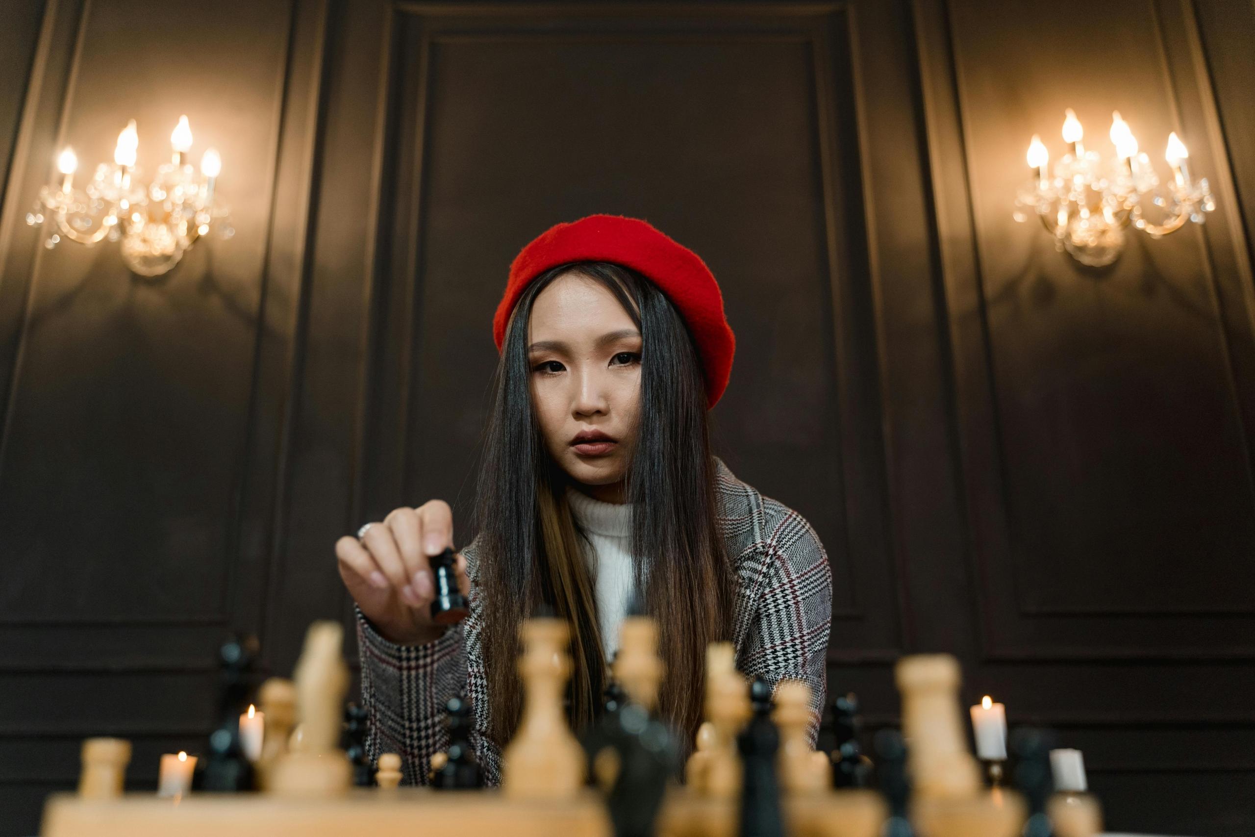 a woman wears a red beret. There are chandeliers lit in the back. She sits in front of a chess board and is about to move a piece.