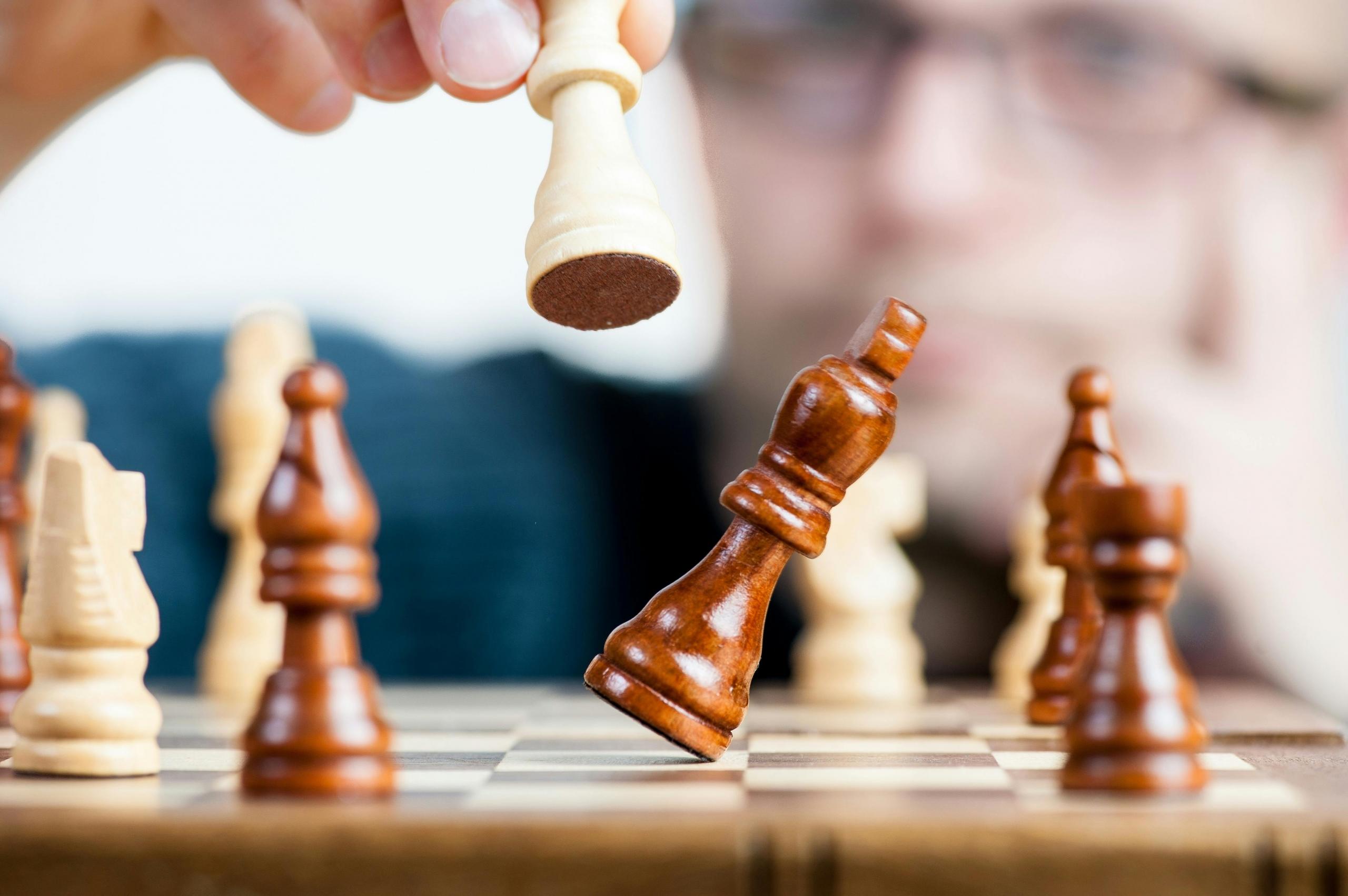 a chess player wearing glasses is seen in the background. He holds a white piece, which knocks over a dark piece.