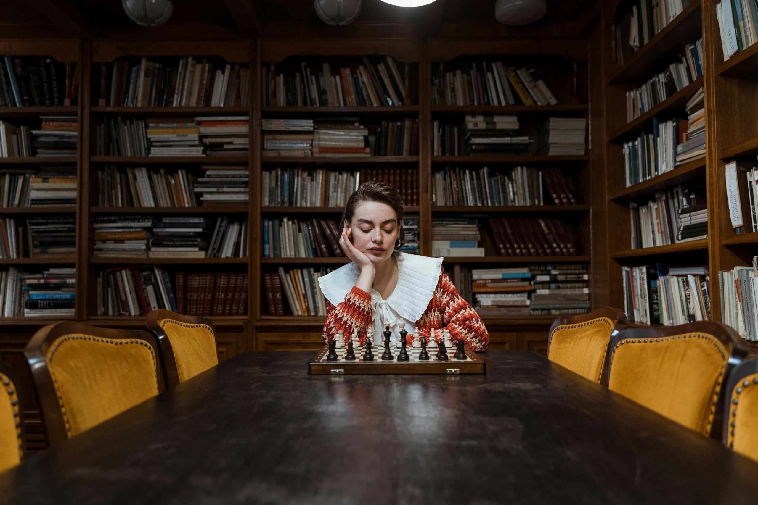 a woman sits in a dark library. A chess board is in front of her. She looks at the board thoughtfully.