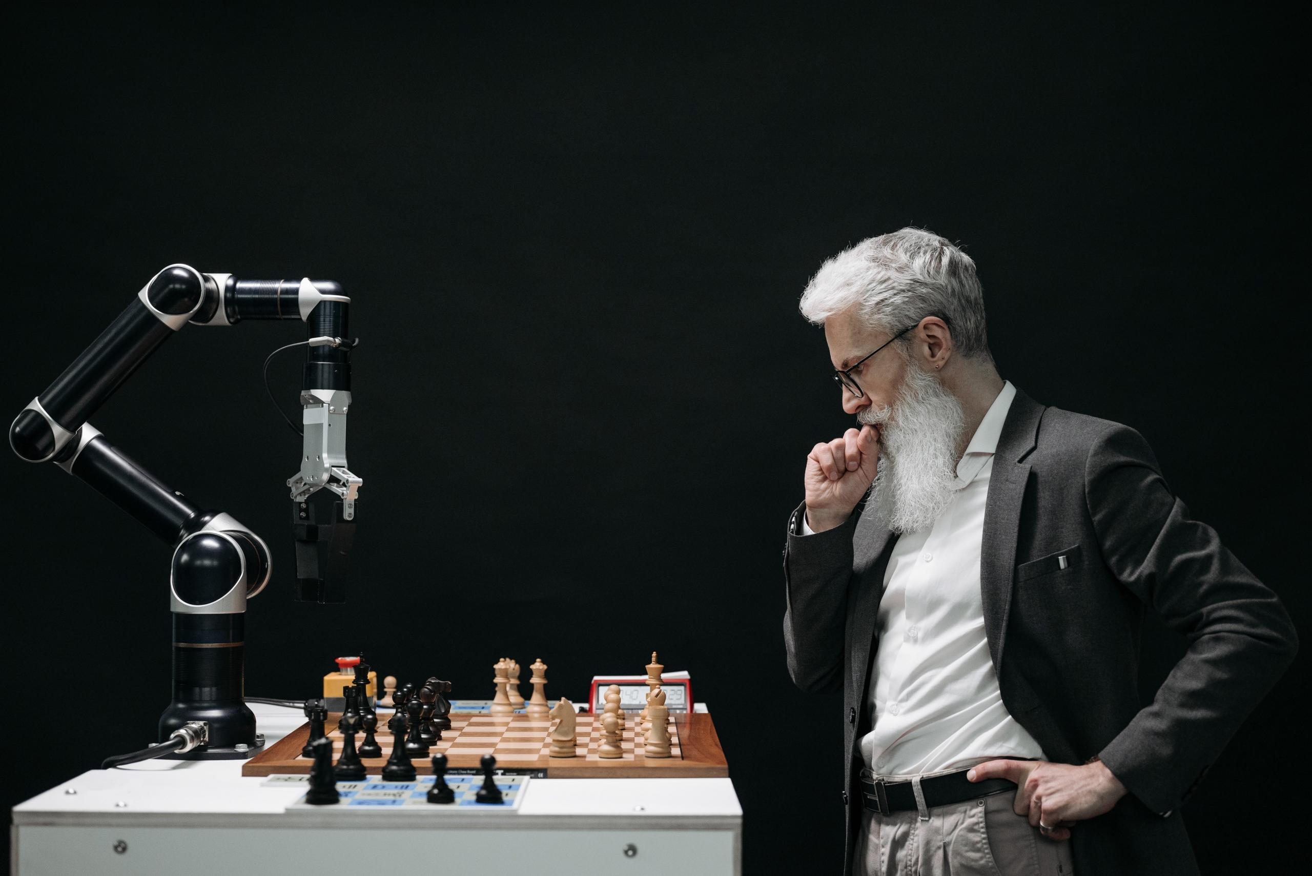 An older man with white hair and a white beard plays chess. A robot is his opponent. The background is dark.