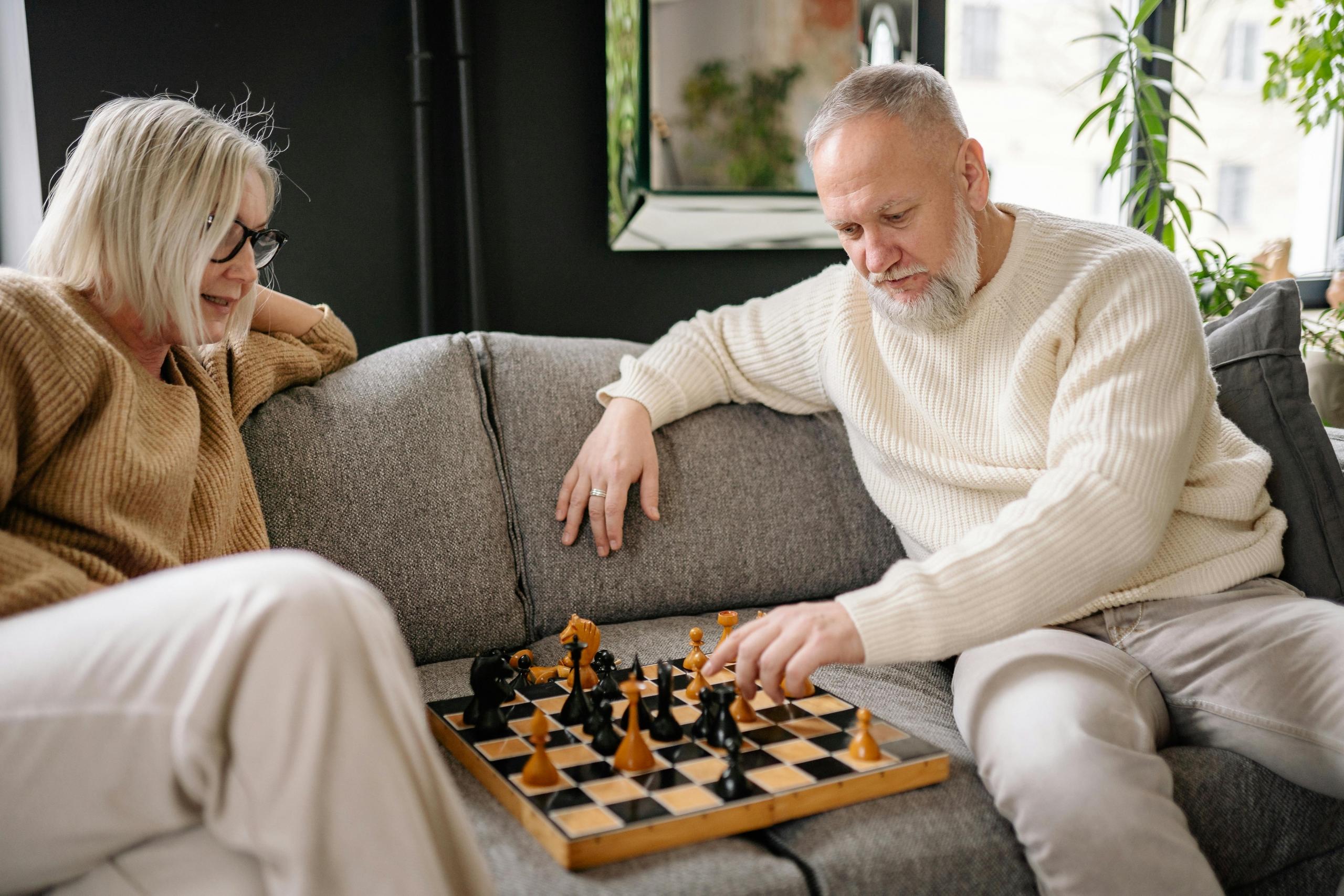 a man and a woman sit on a gray couch. They play chess together. The chess board is on the cushions.