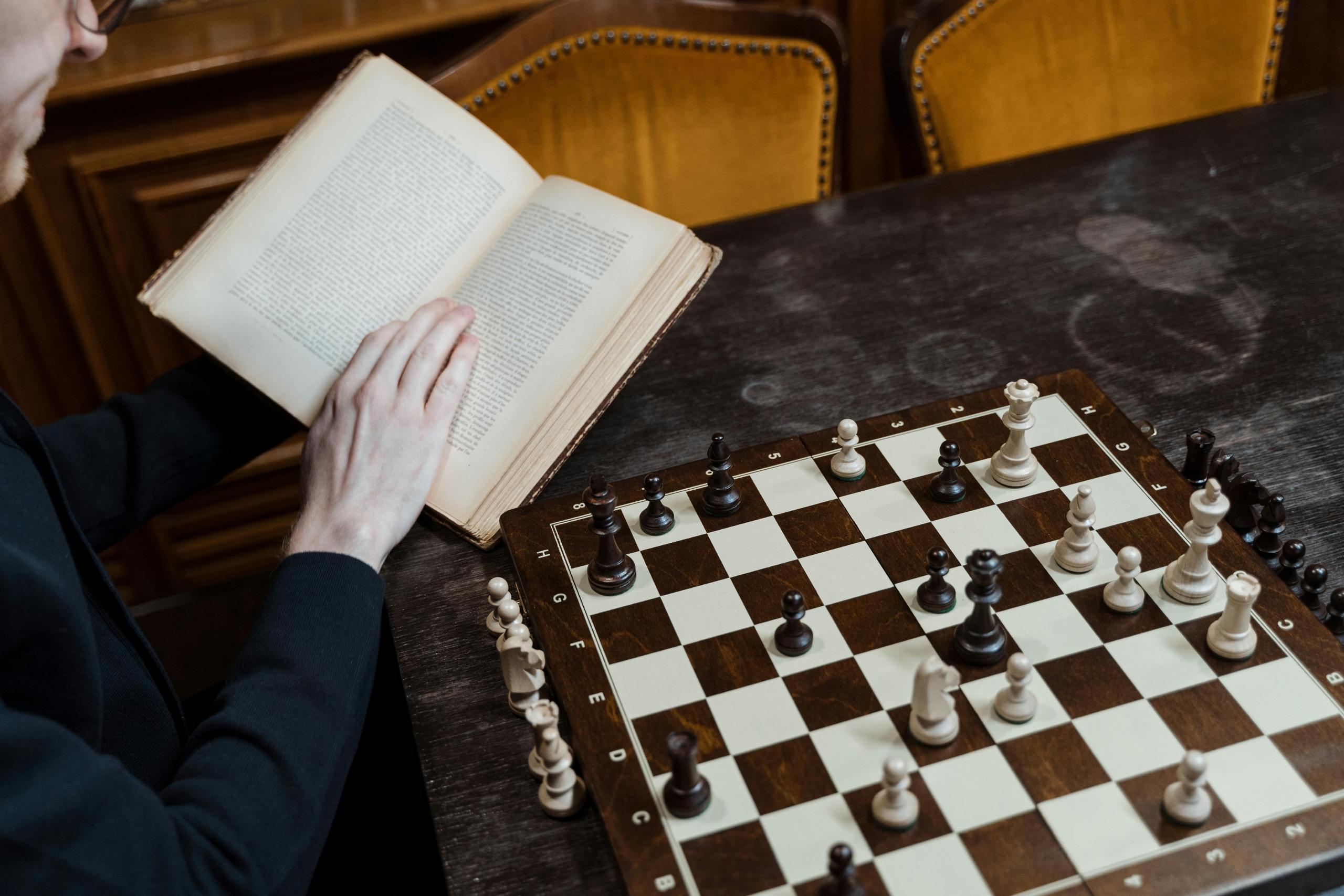 a man reads a book while playing chess. He does not have an opponent.
