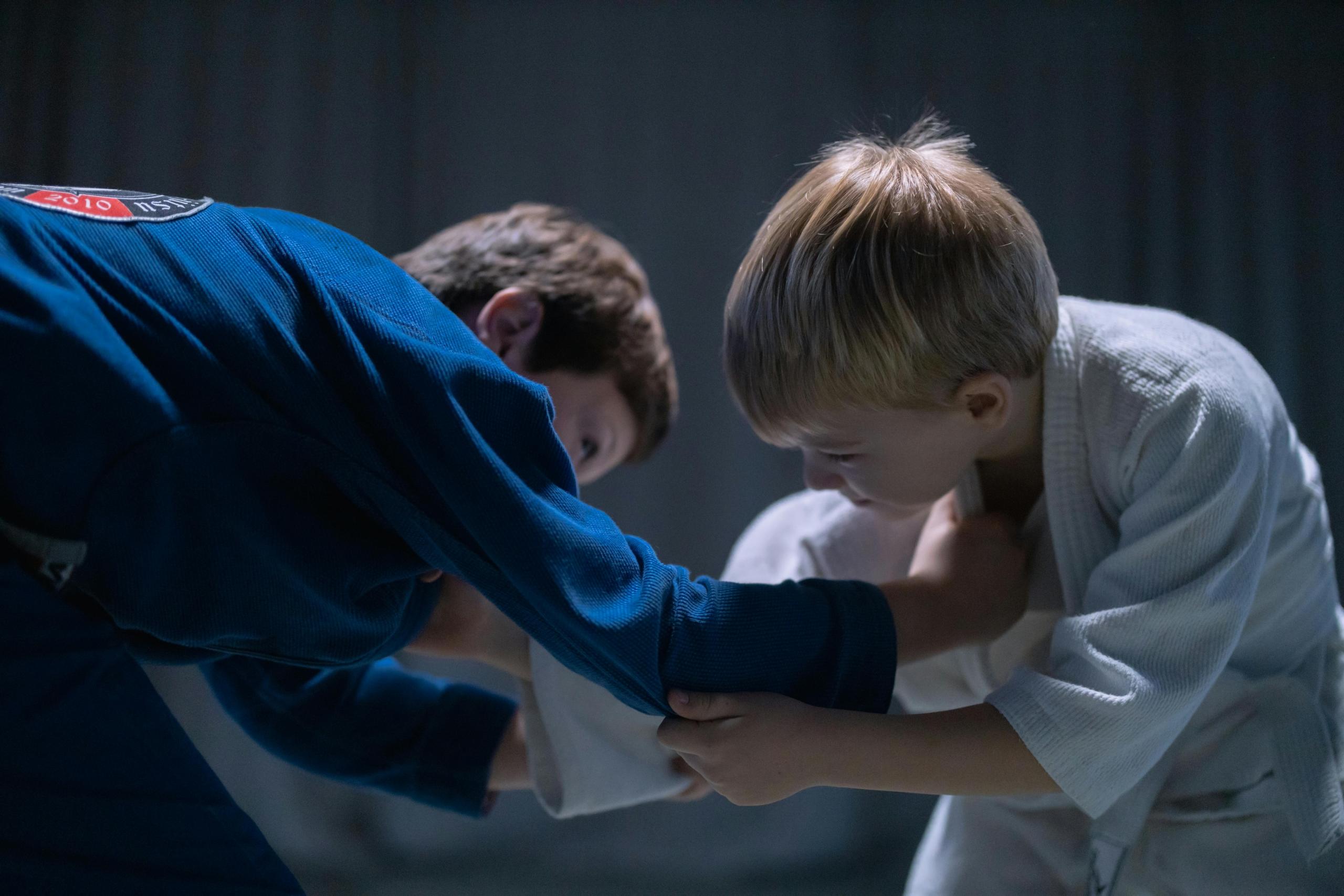 Two young boys practicing Judo on a mat. The boy on the left is wearing a blue judogi, and the boy on the right is wearing a white judogi. They are engaged in a grappling exercise, focusing intensely on each other's movements.