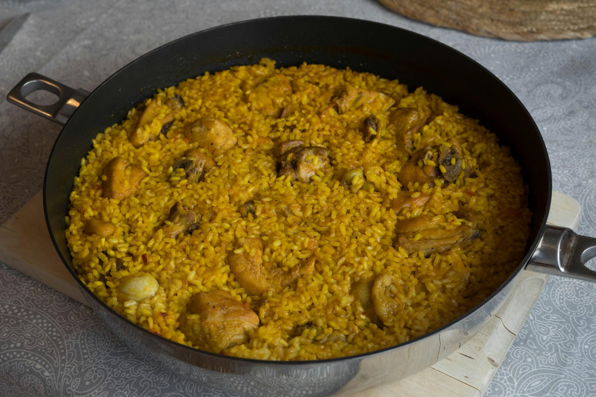 A round pan filled with yellow rice and pieces of chicken, garnished with spices, resting on a wooden surface with a patterned tablecloth.