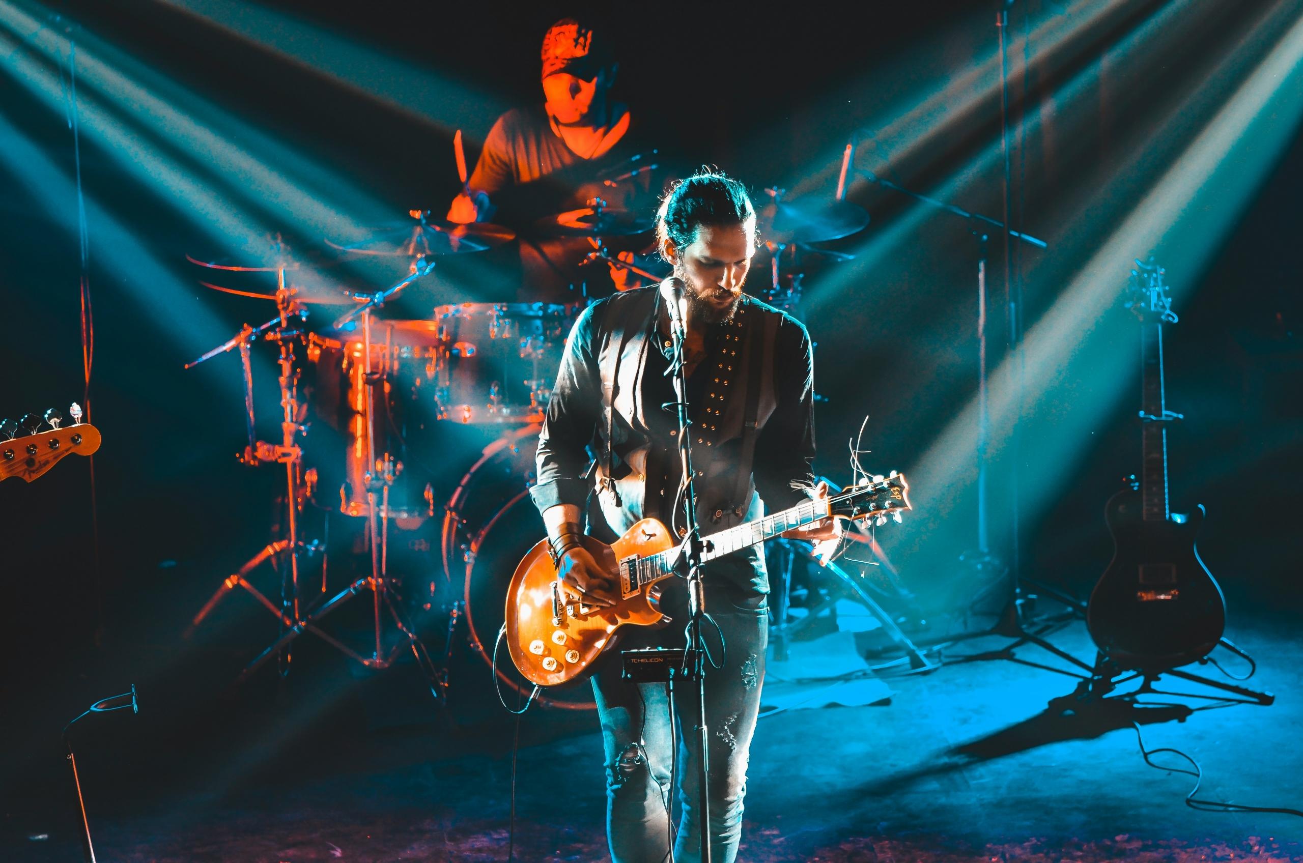 A man playing guitar on stage with a drummer in the background.