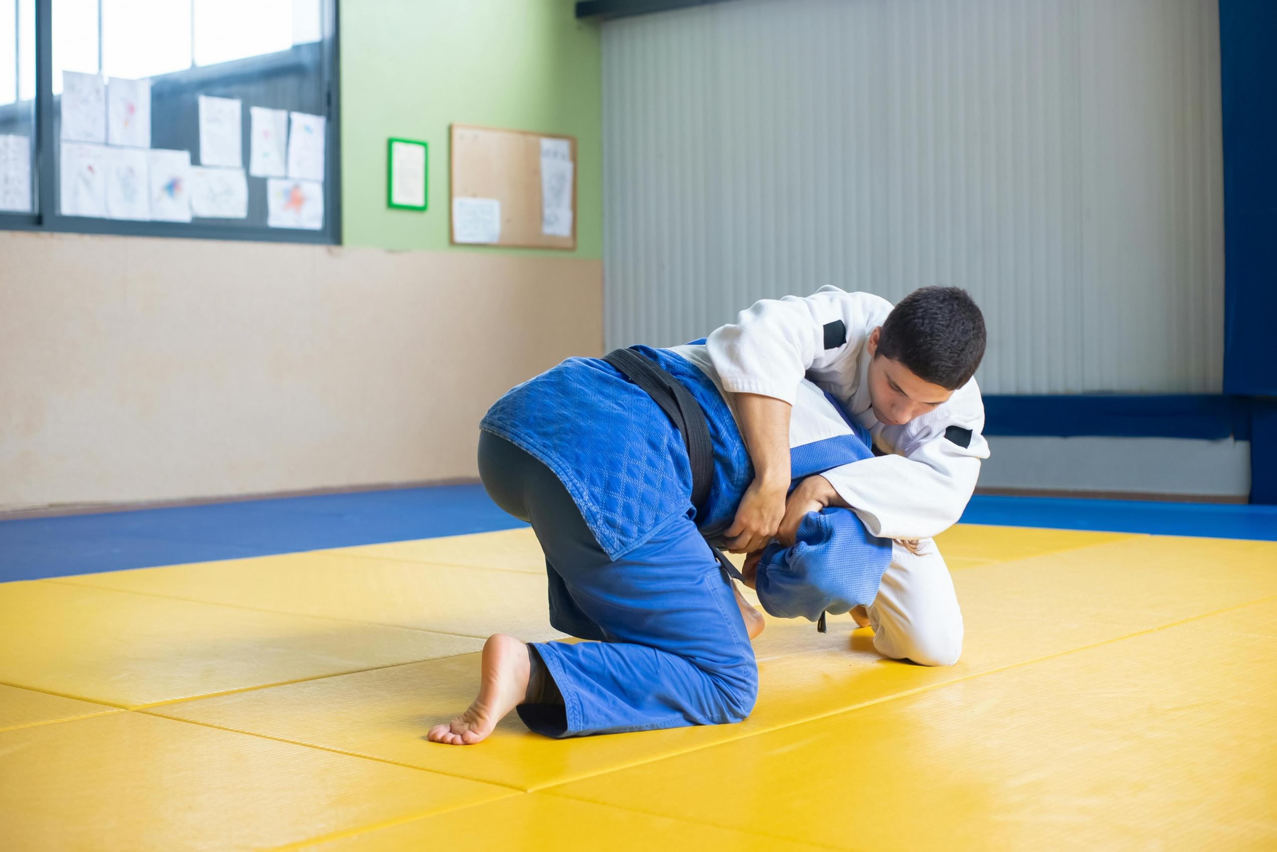 Two male judo practitioners are engaged in a grappling technique on a yellow mat inside a dojo. Both are wearing traditional judo gis, with one in a blue gi and the other in a white gi.