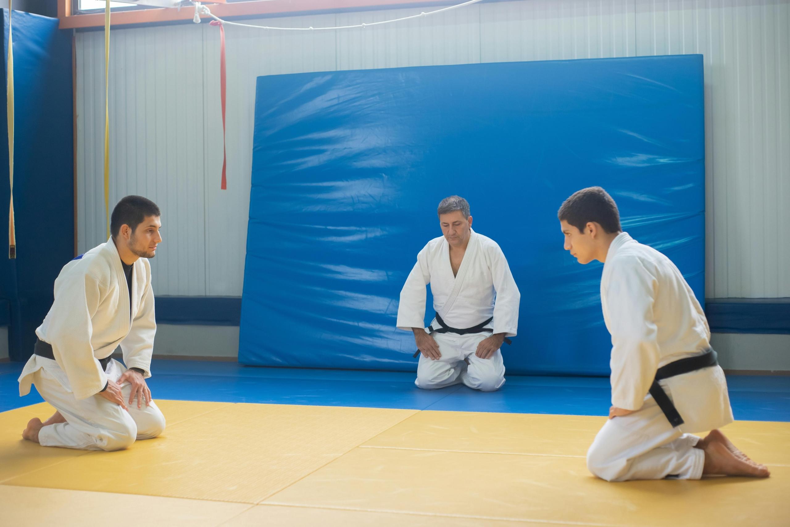 Two Judo practitioners kneel on a yellow mat, facing each other in a dojo, while their instructor, dressed in a white gi, observes in the background. The scene is set for a formal training session, emphasizing discipline and respect in the martial art.