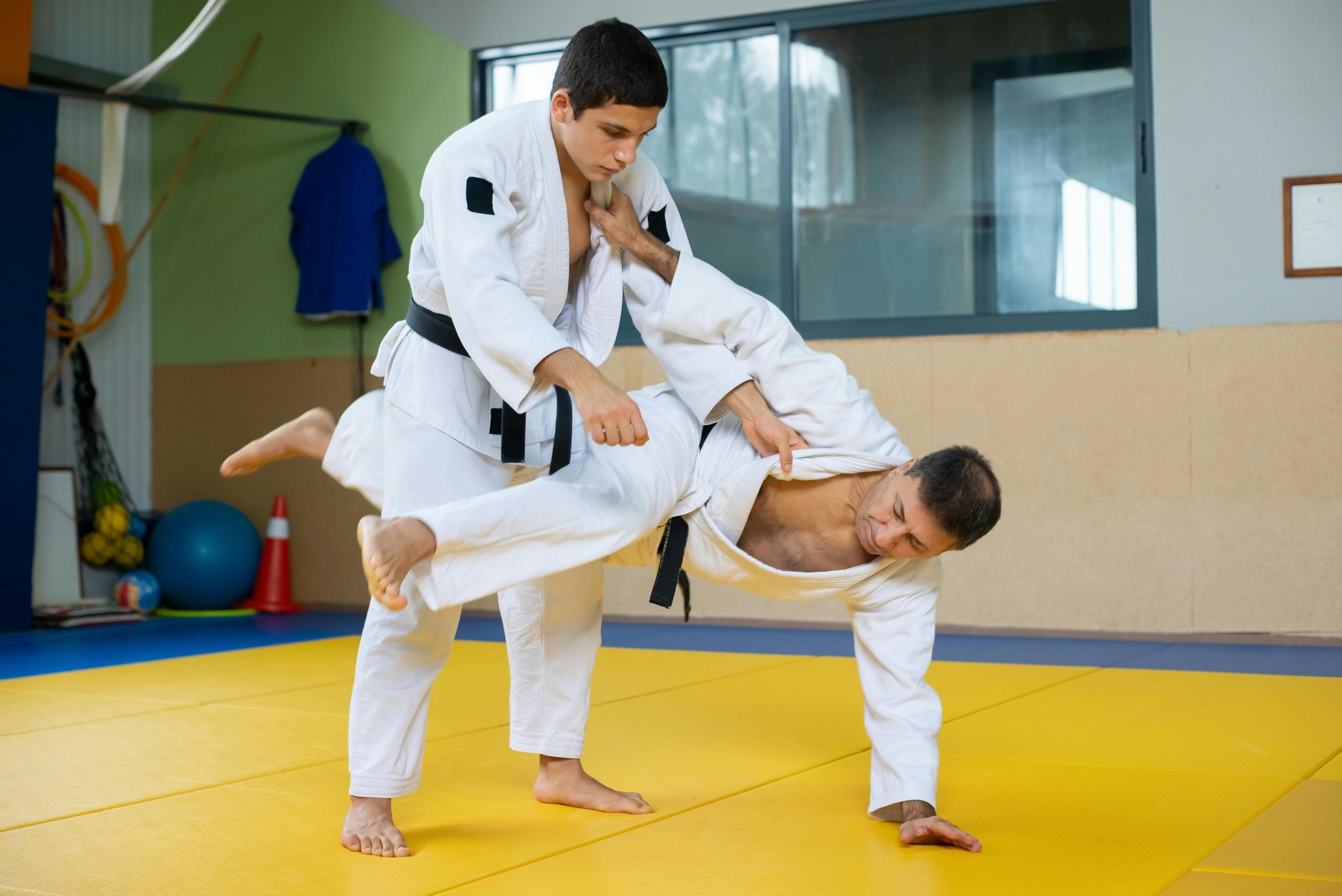Two judokas practice a throw technique on a yellow mat inside a dojo. 