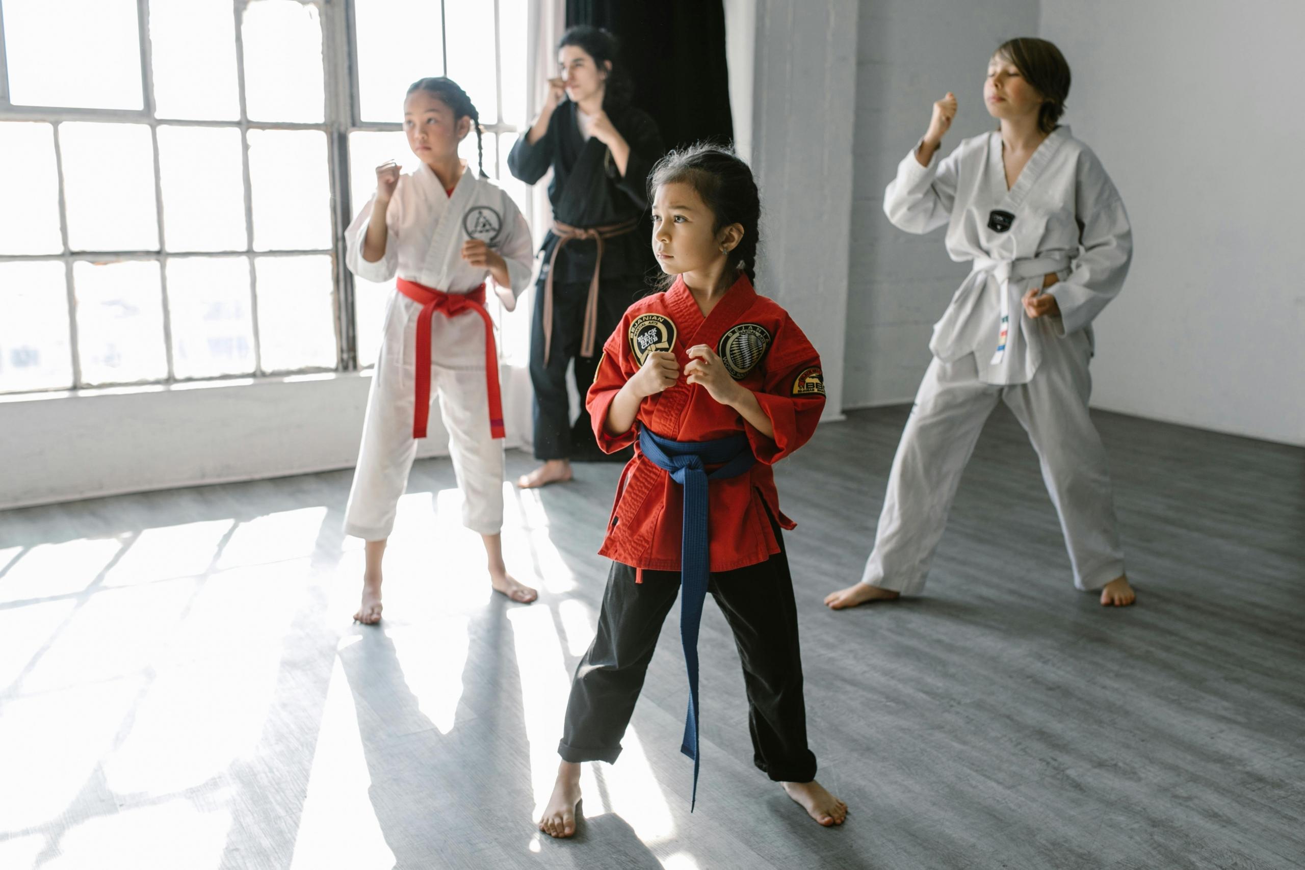 A group of children and an adult instructor practice martial arts in a brightly lit dojo. The children, wearing red and white martial arts uniforms with colored belts, are in fighting stances, focusing intently on their movements. The instructor, dressed in a black uniform, guides them through their techniques. The large windows in the background let in natural light, creating a warm and inviting training environment.