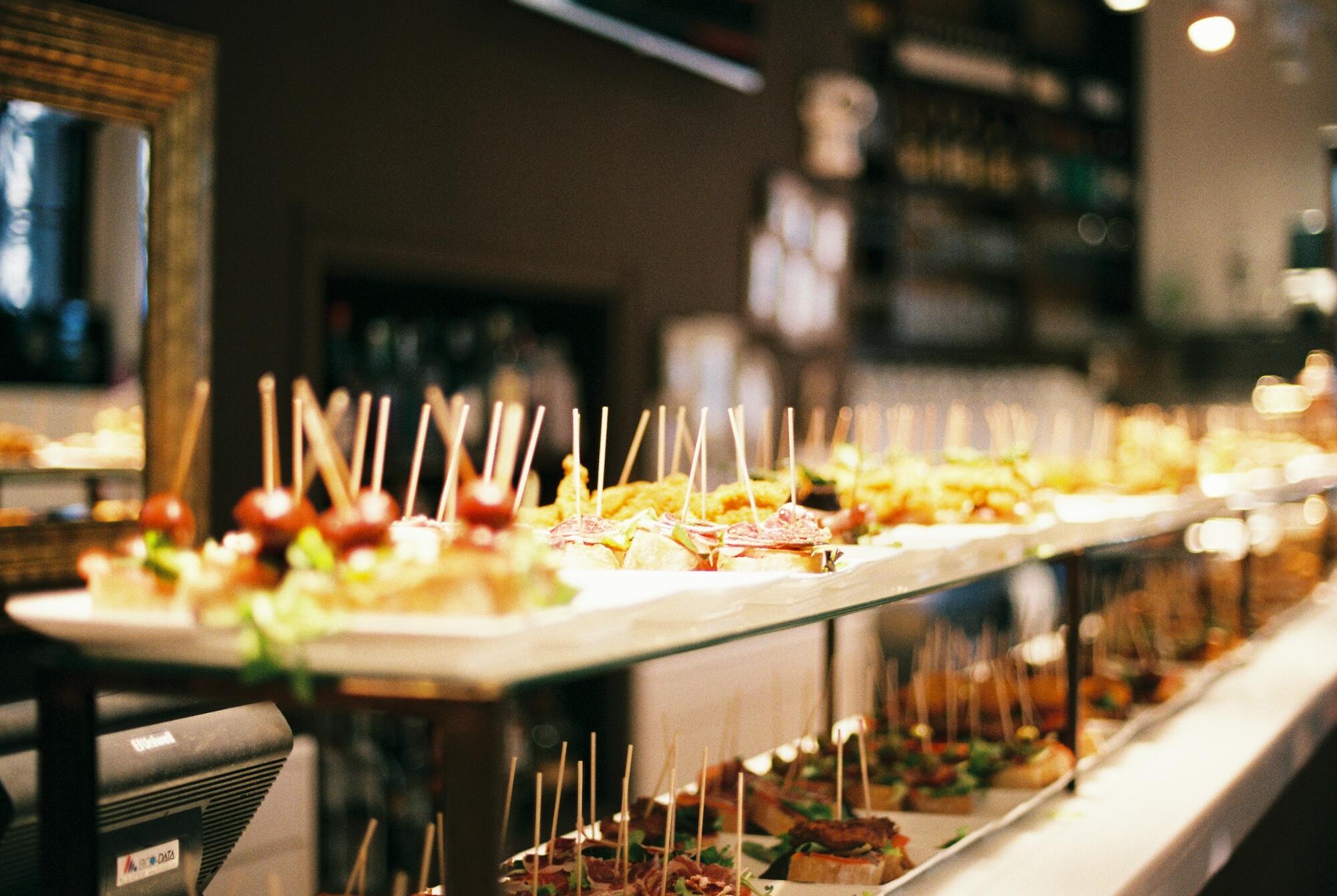 A variety of colorful tapas displayed on plates, garnished with greens and topped with skewers, in a stylish restaurant setting.