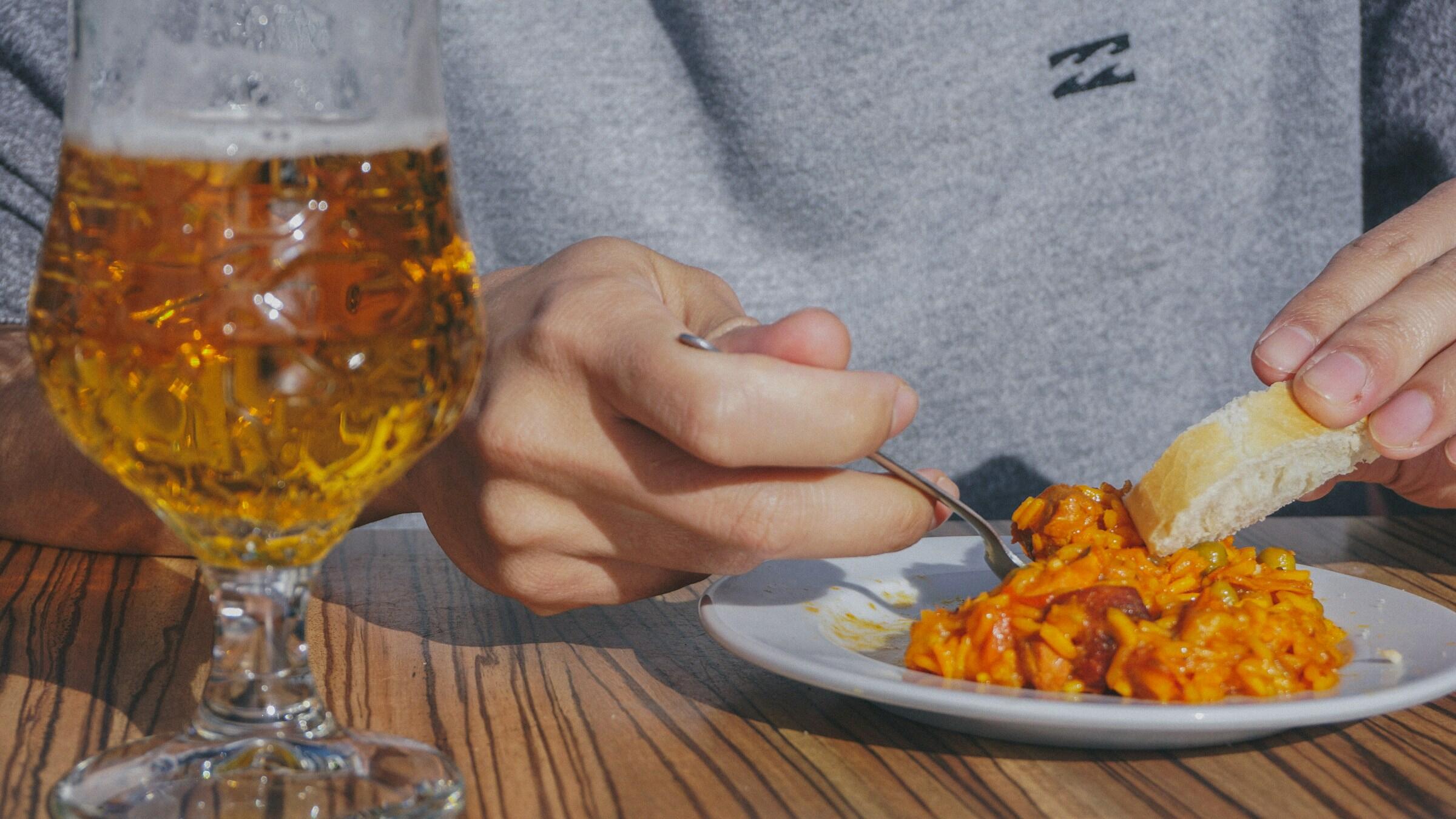 A person enjoys a plate of flavorful rice with a fork, paired with a glass of beer, seated at a wooden table.