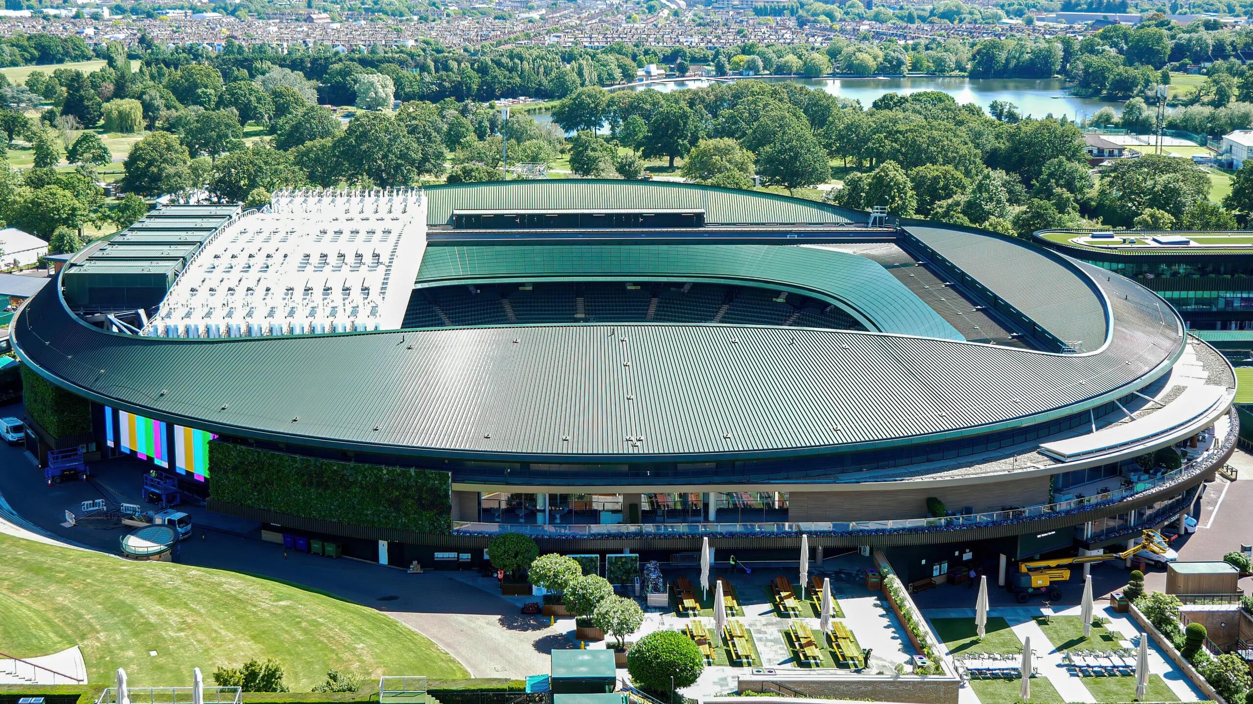 erial view of Wimbledon Stadium showing the iconic Centre Court surrounded by lush green lawns, outer courts.