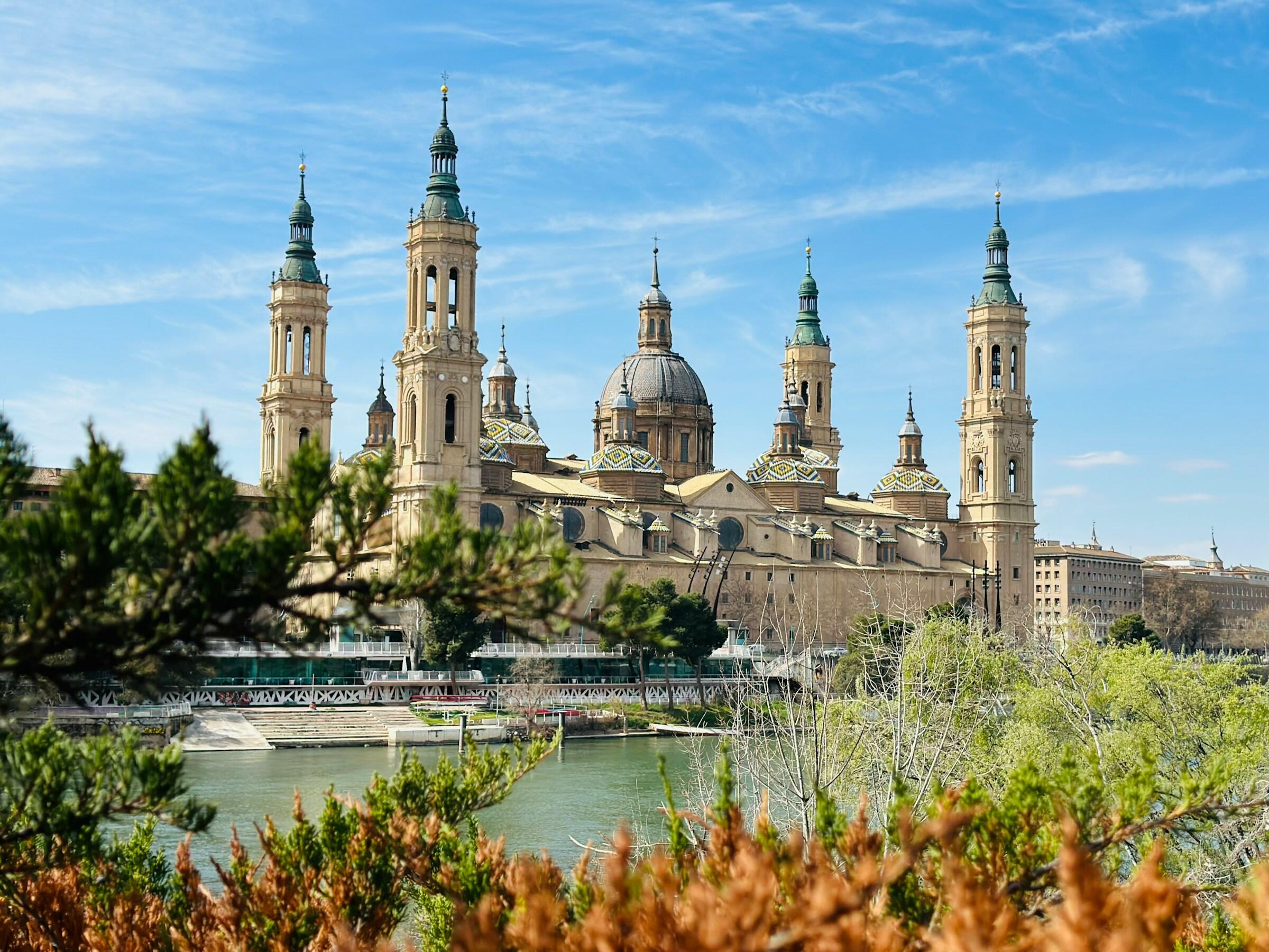 A picturesque view of the Basilica del Pilar in Zaragoza, featuring its distinctive towers and domes against a blue sky.