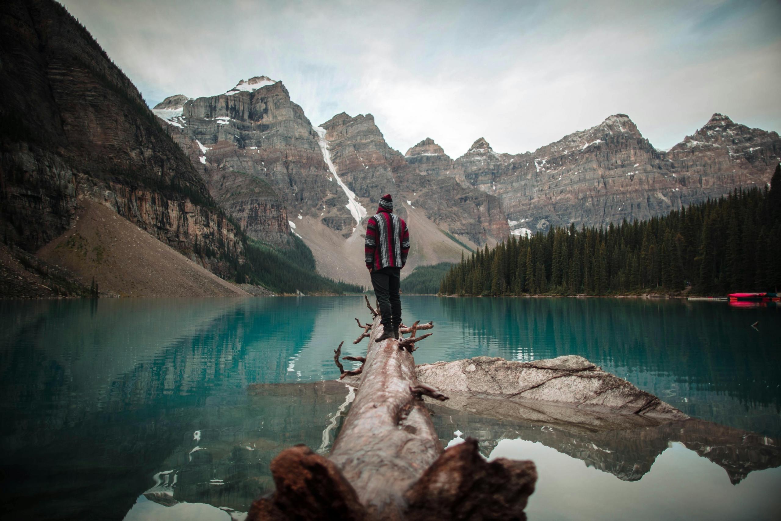 A person stands on a fallen log extending into a clear, turquoise lake surrounded by towering, mountains. The individual is dressed warmly in a red and gray striped sweater and a beanie, gazing at the majestic landscape of tall trees and rocky peaks, with patches of snow.