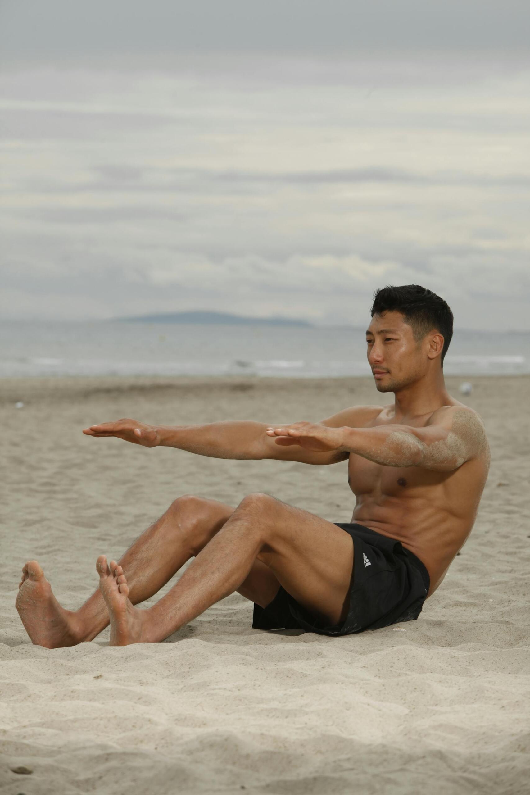 A person sits on the beach, performing a core exercise with legs extended and arms outstretched, against a cloudy sky background.