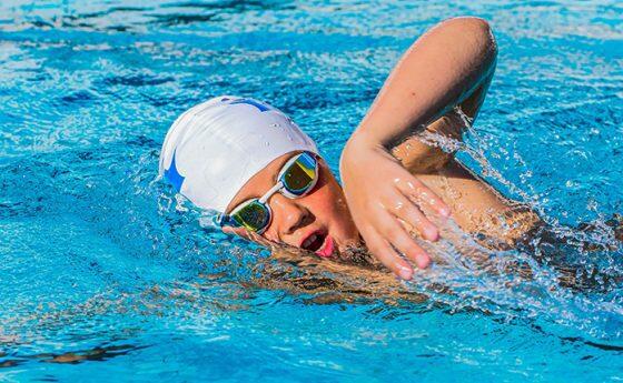 A swimmer glides through clear blue water, creating splashes with their arms in an active swimming posture.
