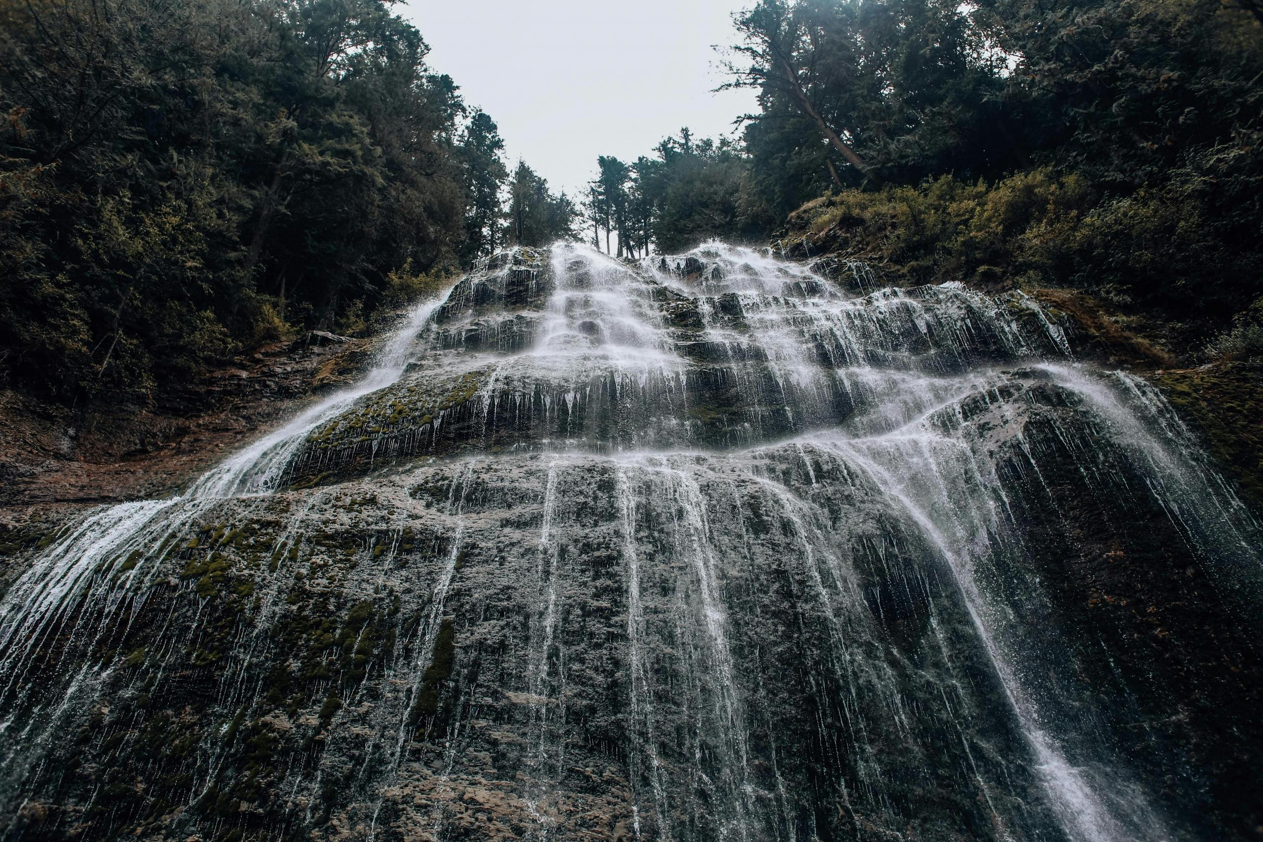 A view of Bridal Veil Falls on Manitoulin Island. The water cascades down a rocky, moss-covered slope surrounded by dense forest. The sky is overcast, adding a serene and natural ambiance to the scene.