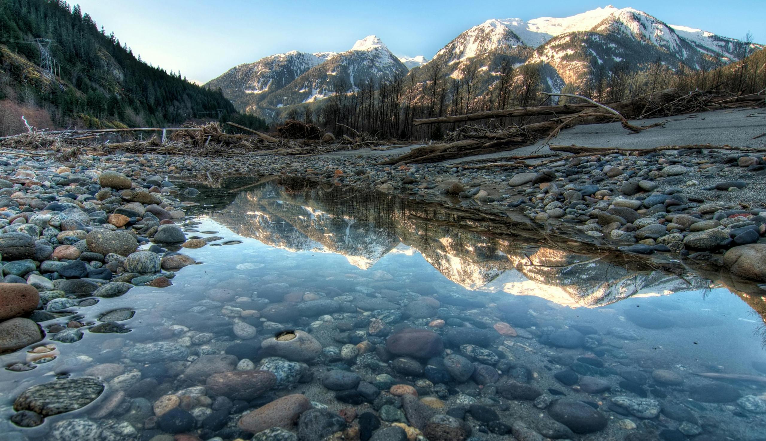 A mountain landscape featuring snow-capped mountains reflected in a calm, shallow water. The foreground is covered with smooth, rounded pebbles and stones of various colors and sizes. Surrounding the pool are fallen logs and branches, with dense, evergreen forests on both sides of the valley.