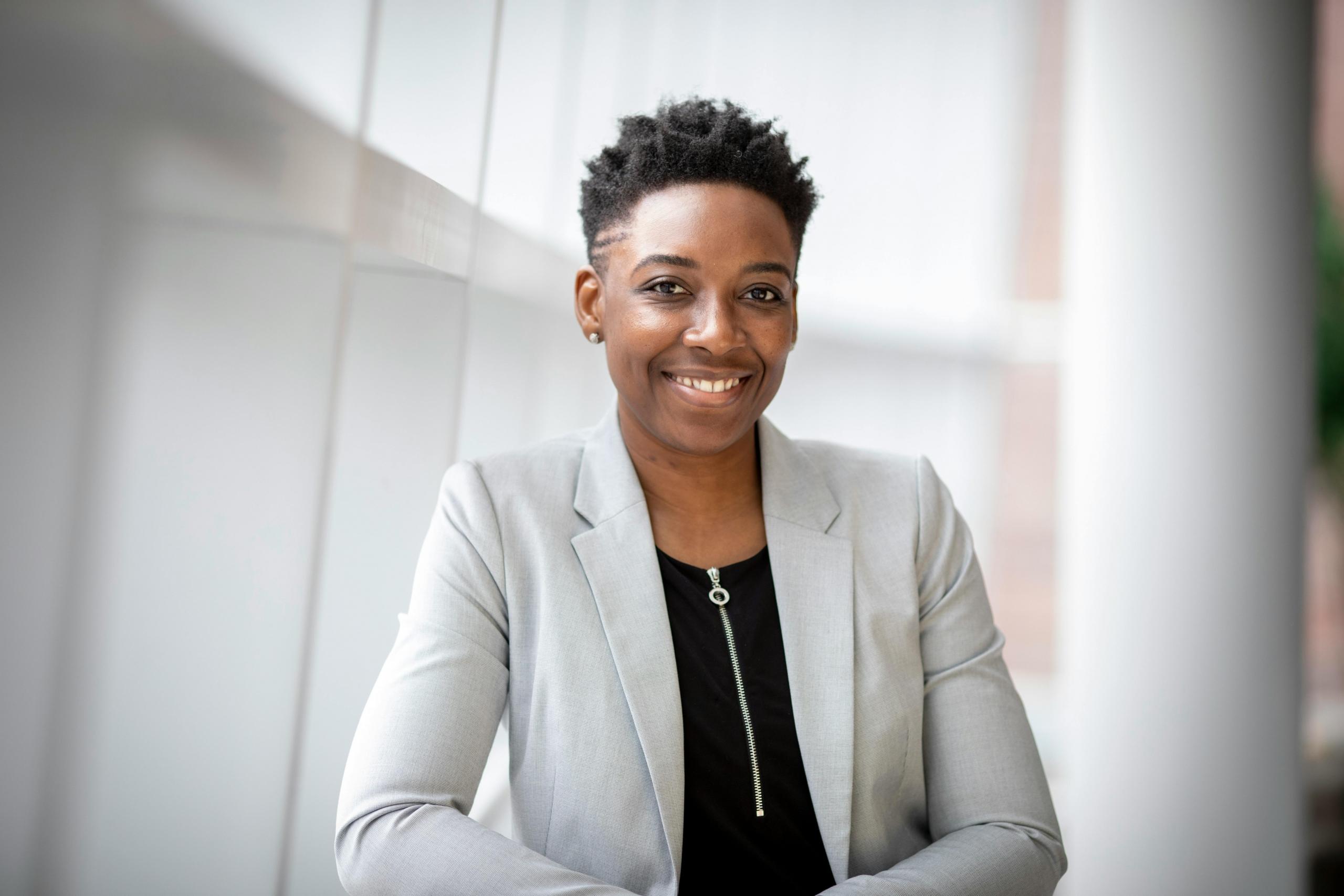 A professional woman in a white blazer smiles at the camera. The background has a white pillar.