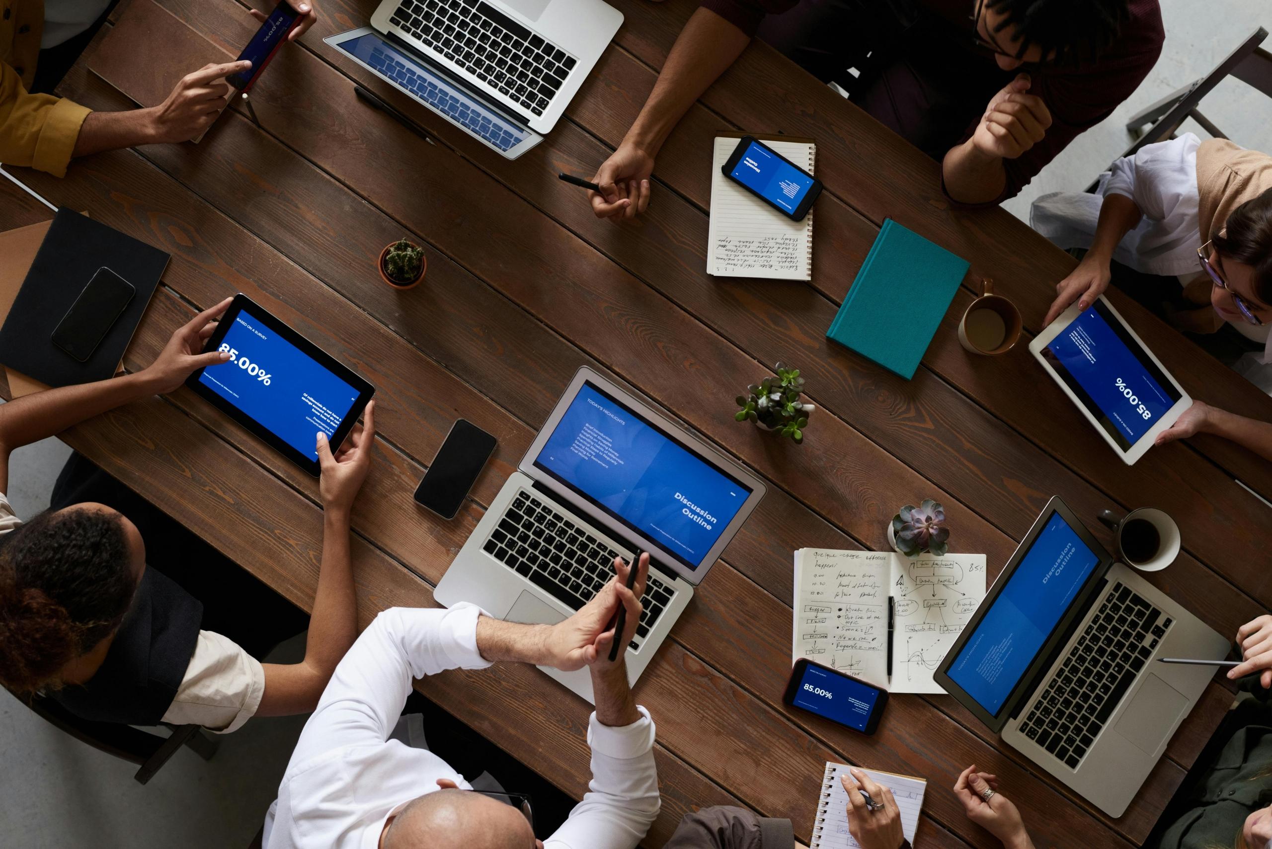 A group of professionals work on laptops. The photo is taken from overhead. The table is brown wood.