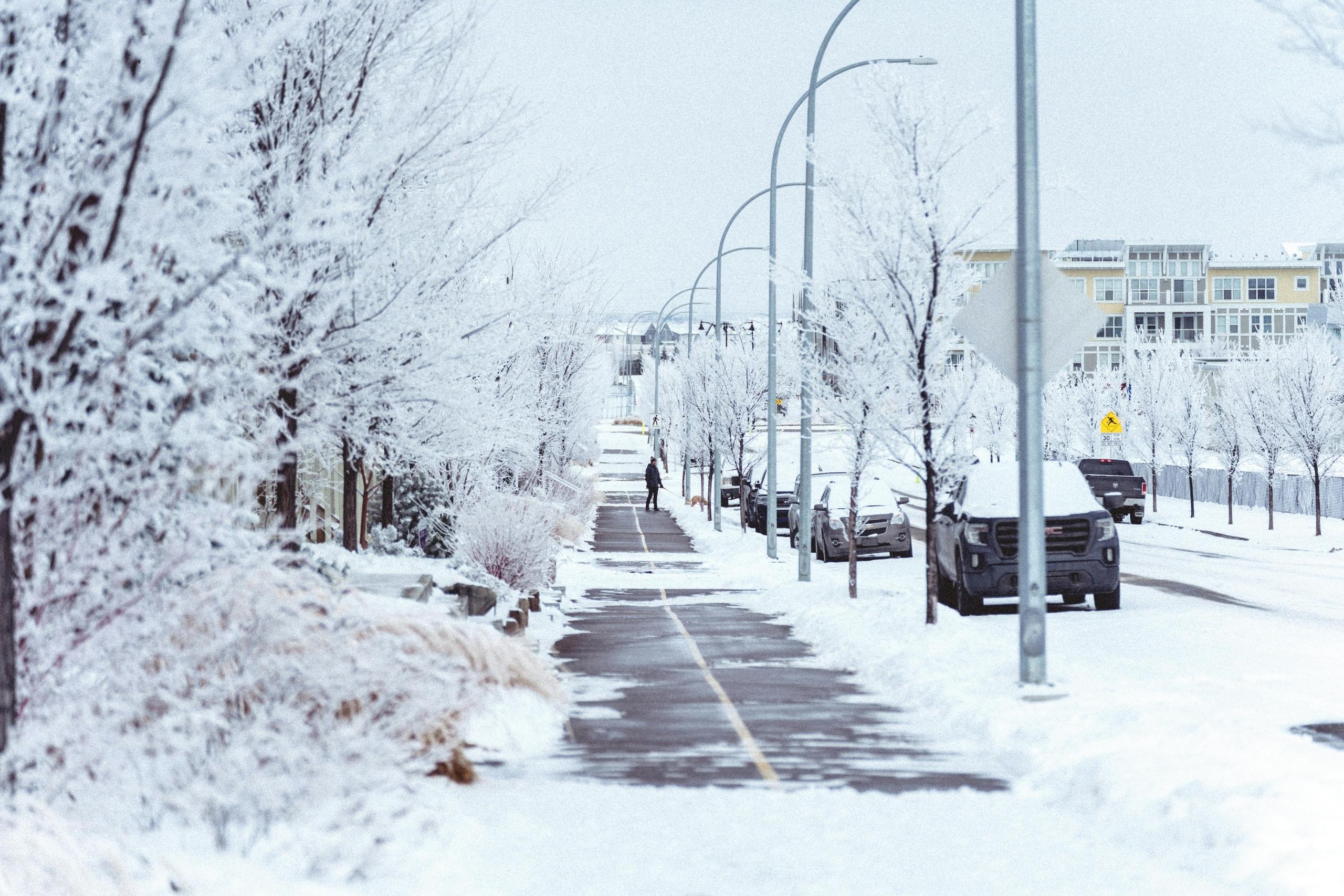 A neighbourhood is covered in snow. A man can be seen on a path beside parked cars. An apartment building is in the background.