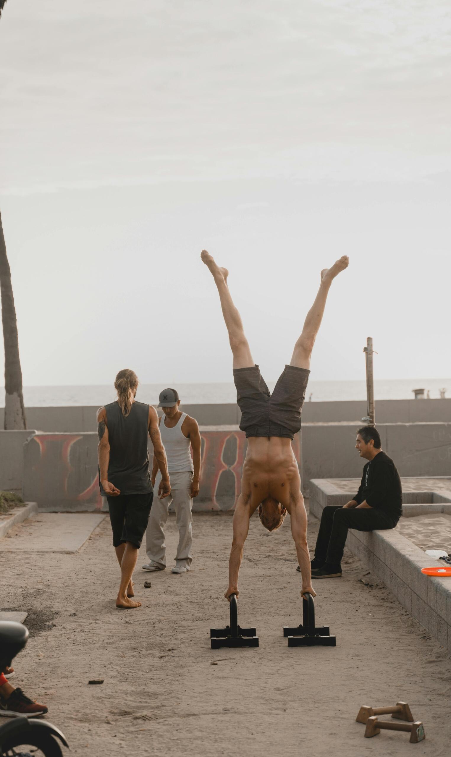 Shirtless man performing a handstand on parallel bars at an outdoor calisthenics park near the beach, surrounded by other people.
