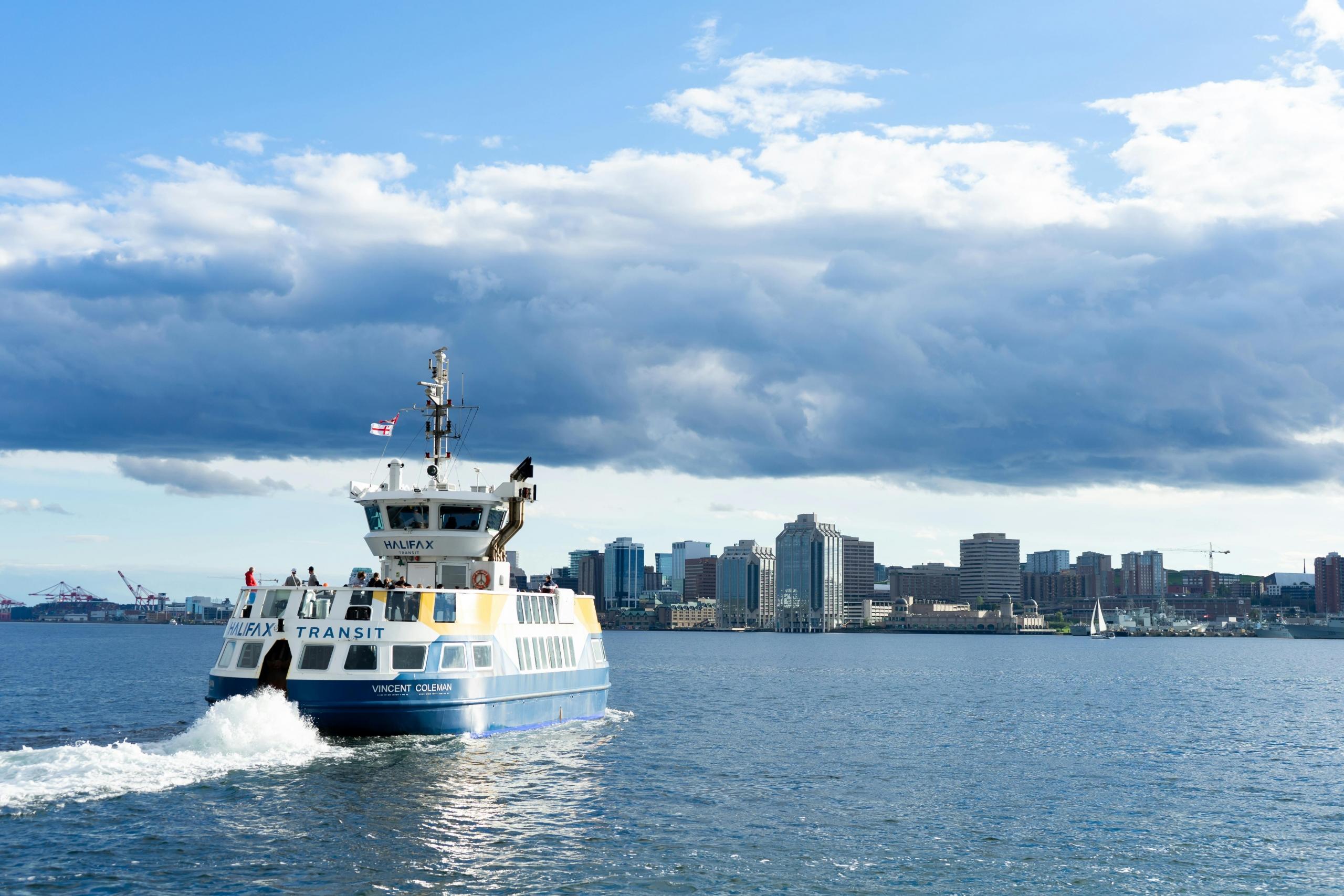 a ferry boat sails toward Halifax. The water is blue and some clouds are overhead.