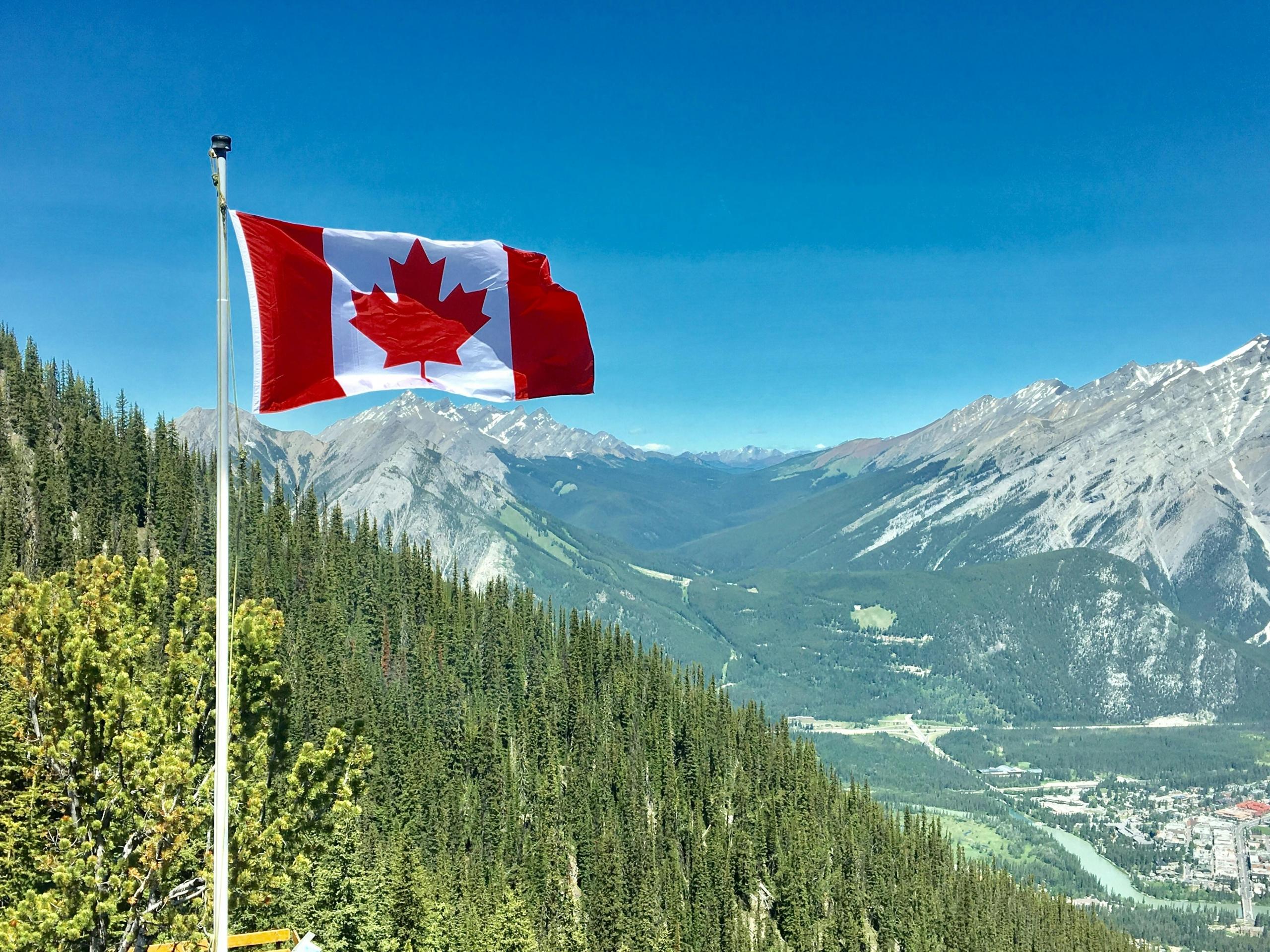 A Canadian flag waves on a flagpole, set against a backdrop of green forests and towering mountains under a clear blue sky.