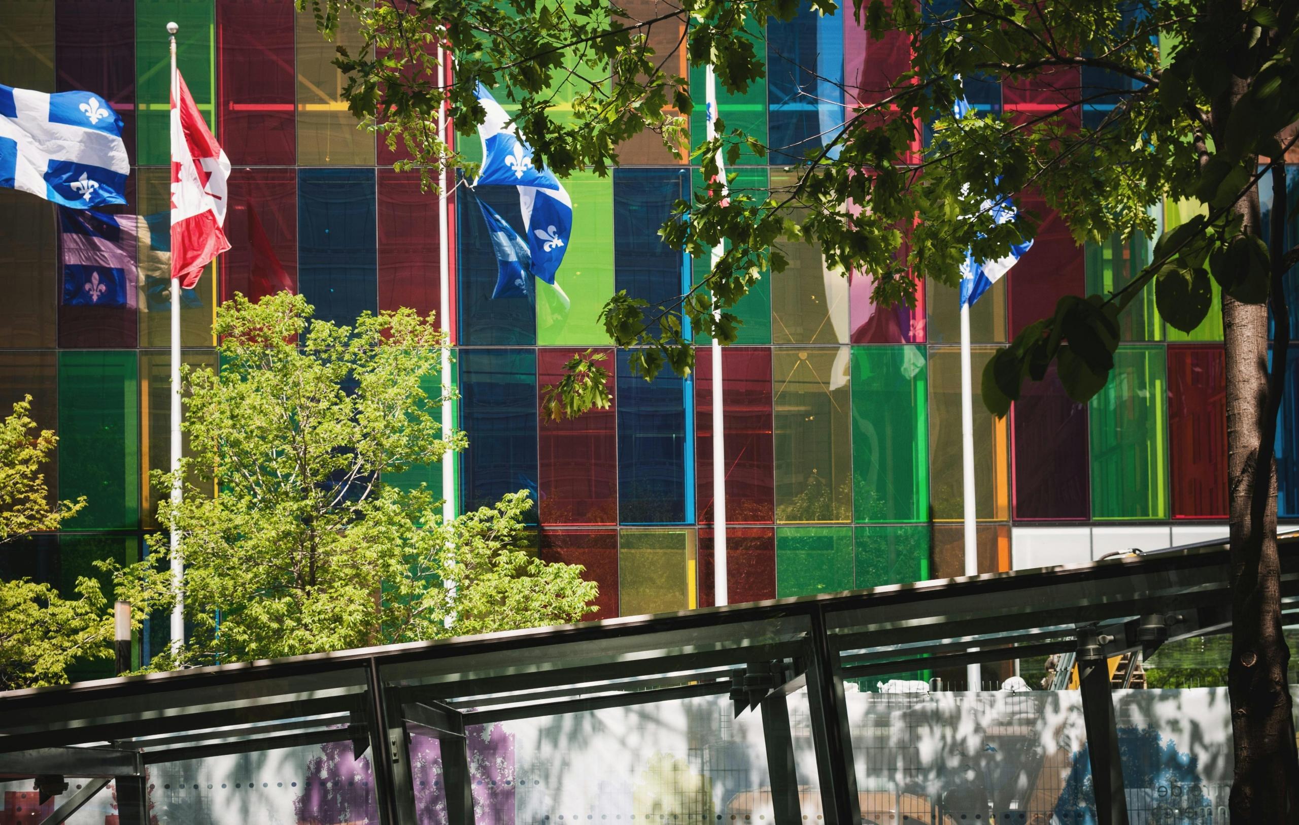 a colourful glass building with multiple flags, including the Quebec flag. The day is sunny.