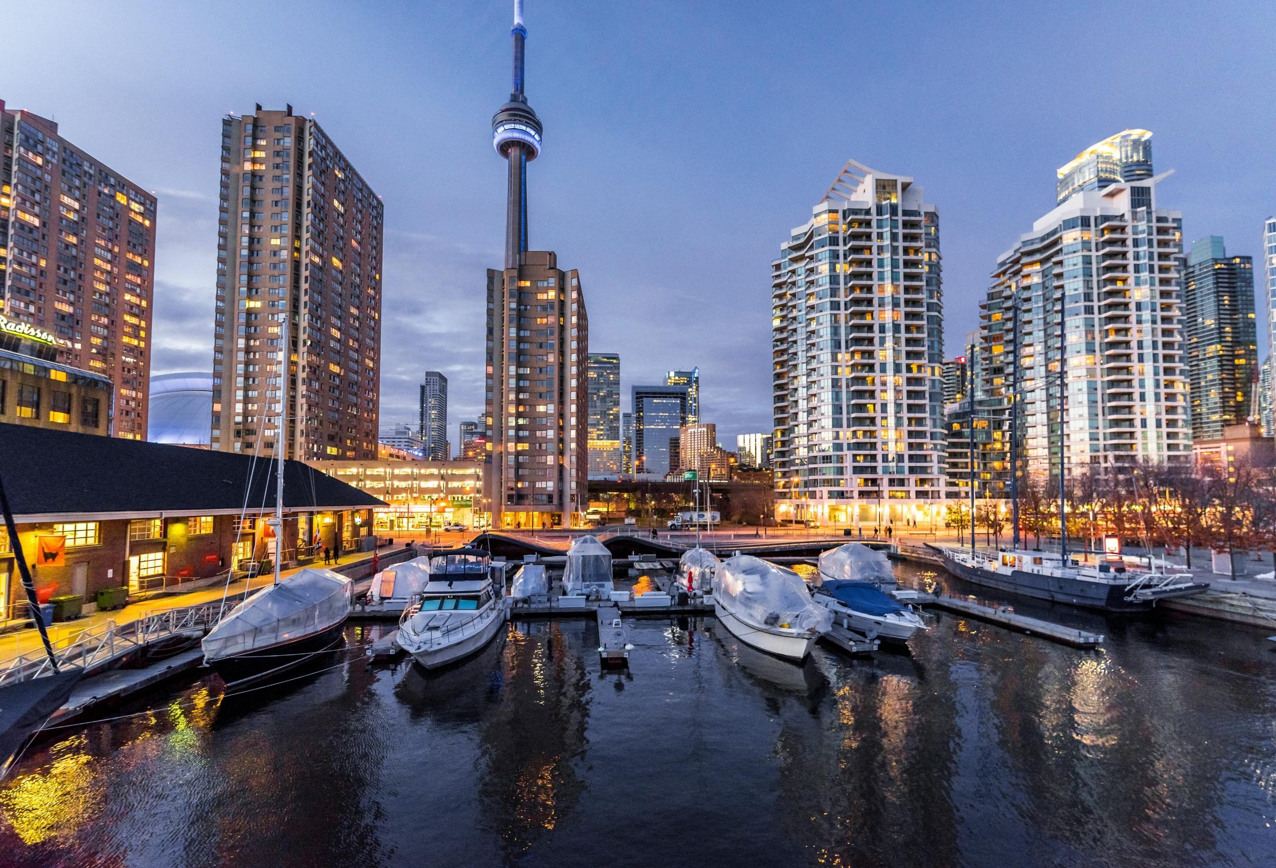 A view of Toronto's city skyline at dusk, featuring the iconic CN Tower surrounded by high-rise buildings. In the foreground, there is a marina with docked boats, reflecting the city lights on the water.
