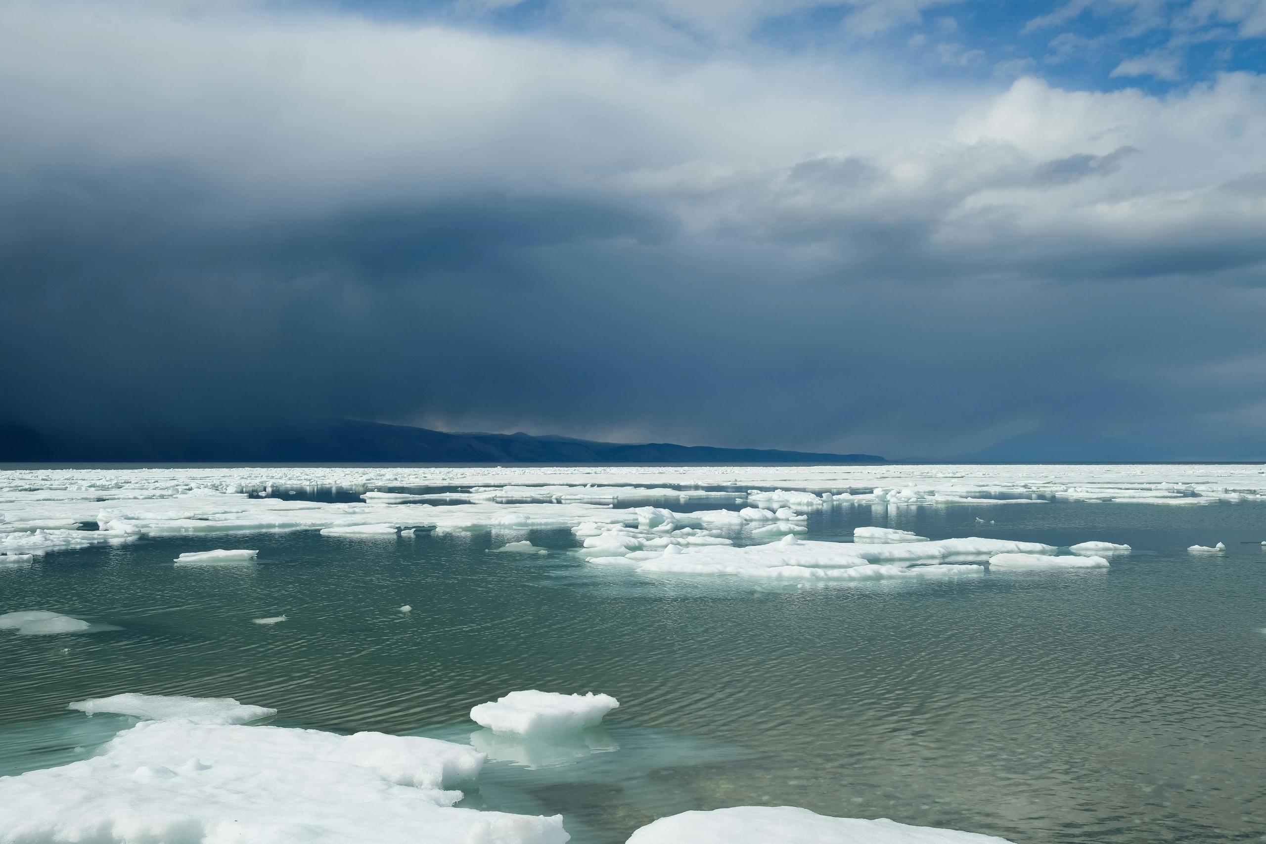 A vast expanse of icy water with chunks of floating sea ice. The sky is partly cloudy with dark clouds on the horizon, and distant mountains are visible in the background.