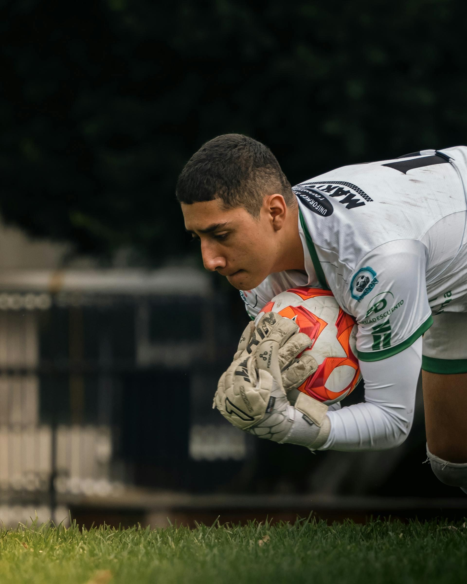 Soccer goalkeeper in white uniform and gloves crouching and holding a red and white ball on a grassy field.