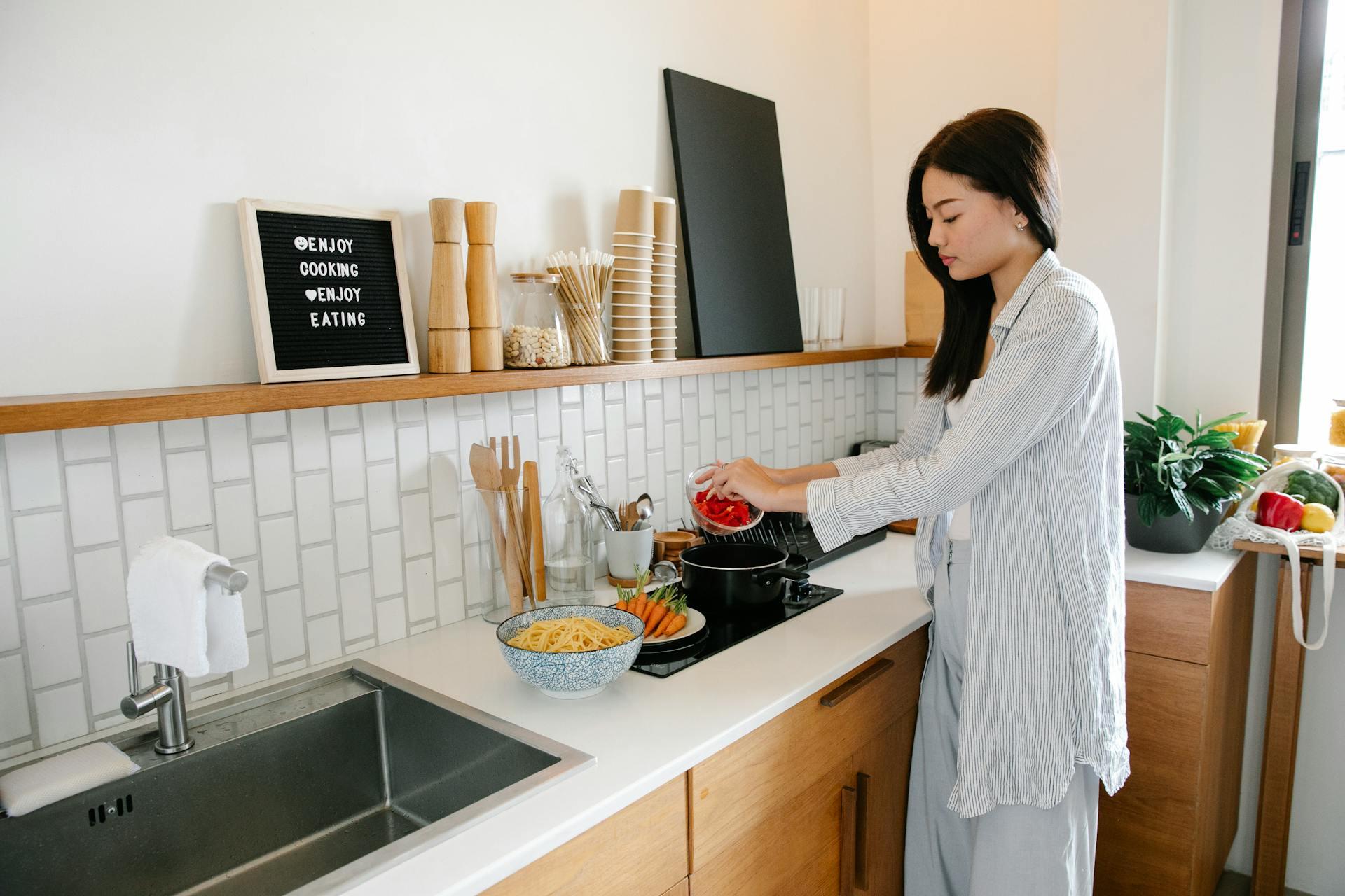 A lady putting red peppers into a pot on the stove while carrots and pasta are beside here. 