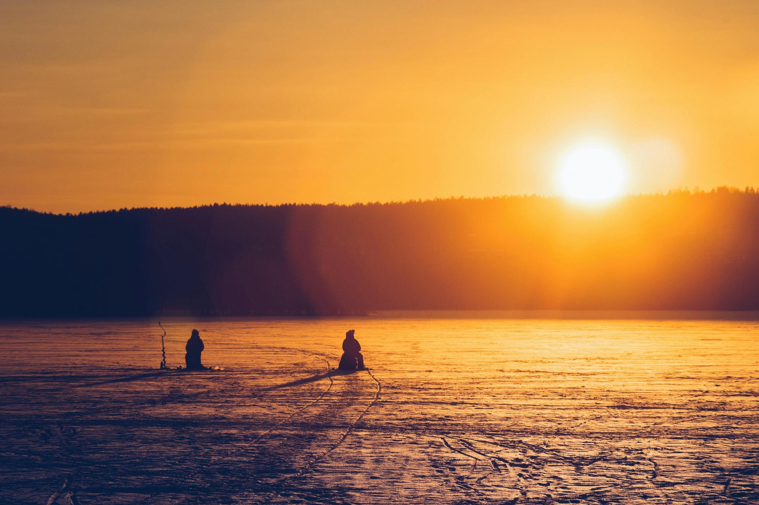 Two people are ice fishing on a frozen lake during a vibrant orange sunset. The silhouettes of the fishers and their equipment are highlighted against the glowing sky, with a forested horizon in the background.