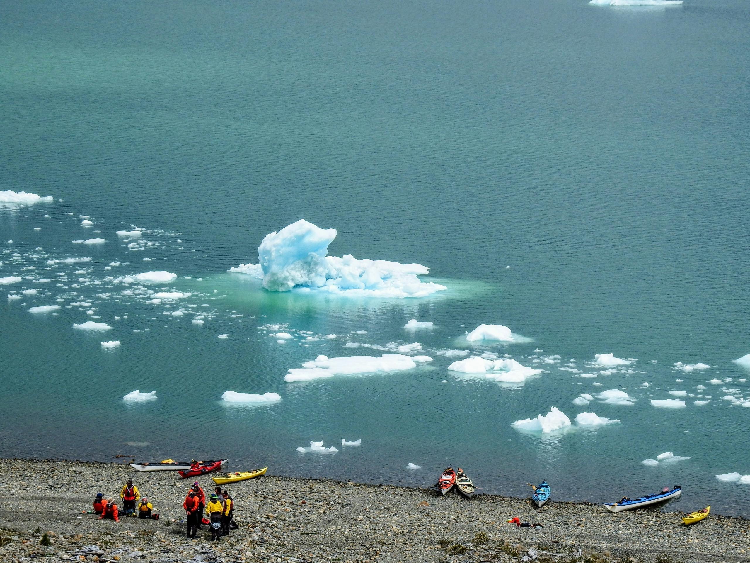A group of kayakers dressed in bright orange and yellow gear prepare their kayaks on a rocky shoreline. In the calm, icy waters nearby, small icebergs and ice chunks float.