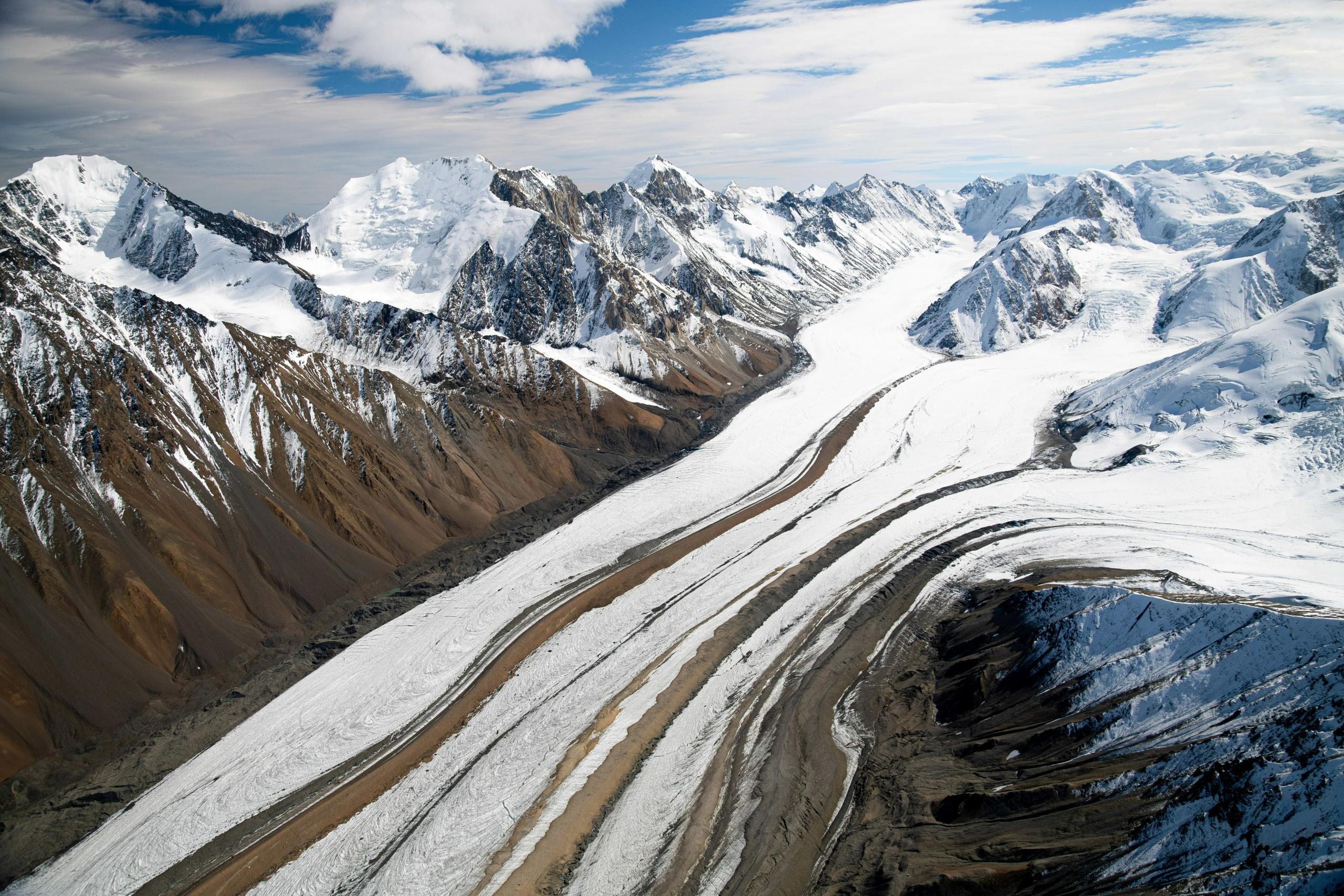 Aerial view of snow-capped mountains, vast glaciers, and rugged terrain under a partly cloudy sky.