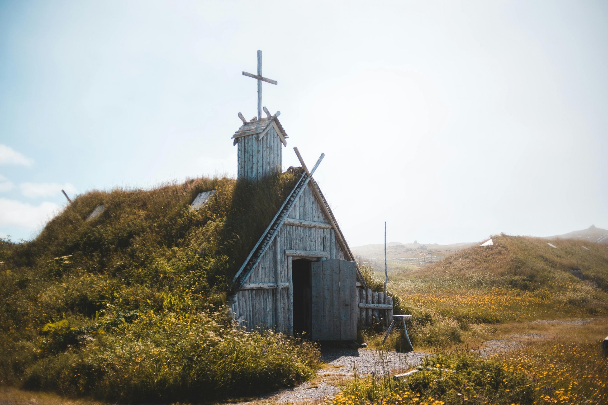 A rustic, sod-roofed wooden structure stands amidst a grassy, flower-filled meadow under a bright, sky. The building features a cross on the roof and wooden planks.