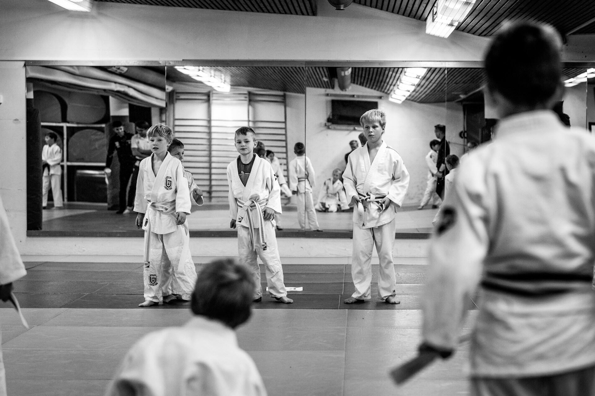 Black and white photo of children in martial arts uniforms standing on mats in a dojo with mirrored walls.