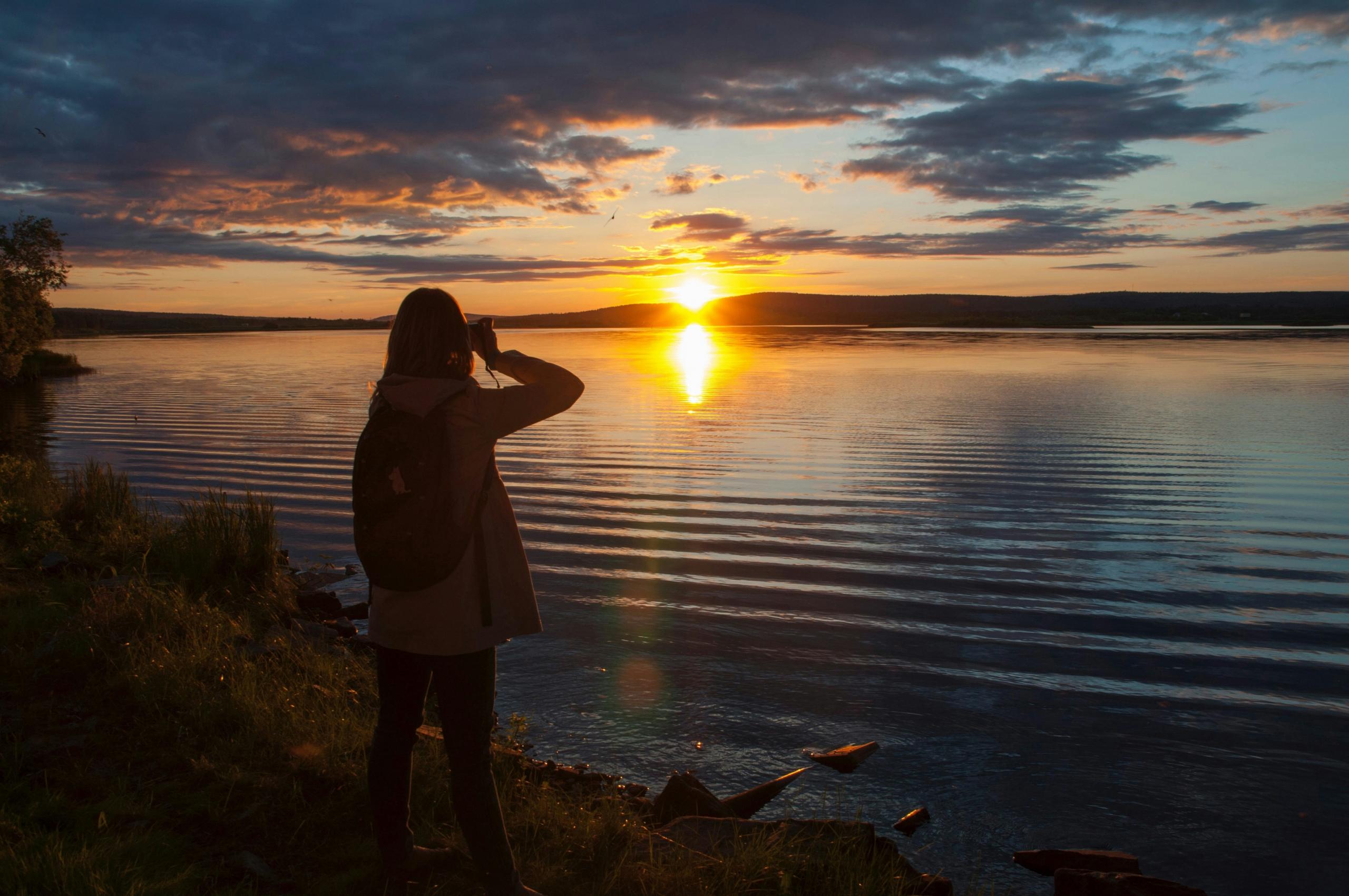 A person stands on the grassy shore of a lake, photographing the midnight sun that casts a golden glow across the water. The sky is filled with dramatic clouds, and the silhouette of the landscape is visible in the distance.