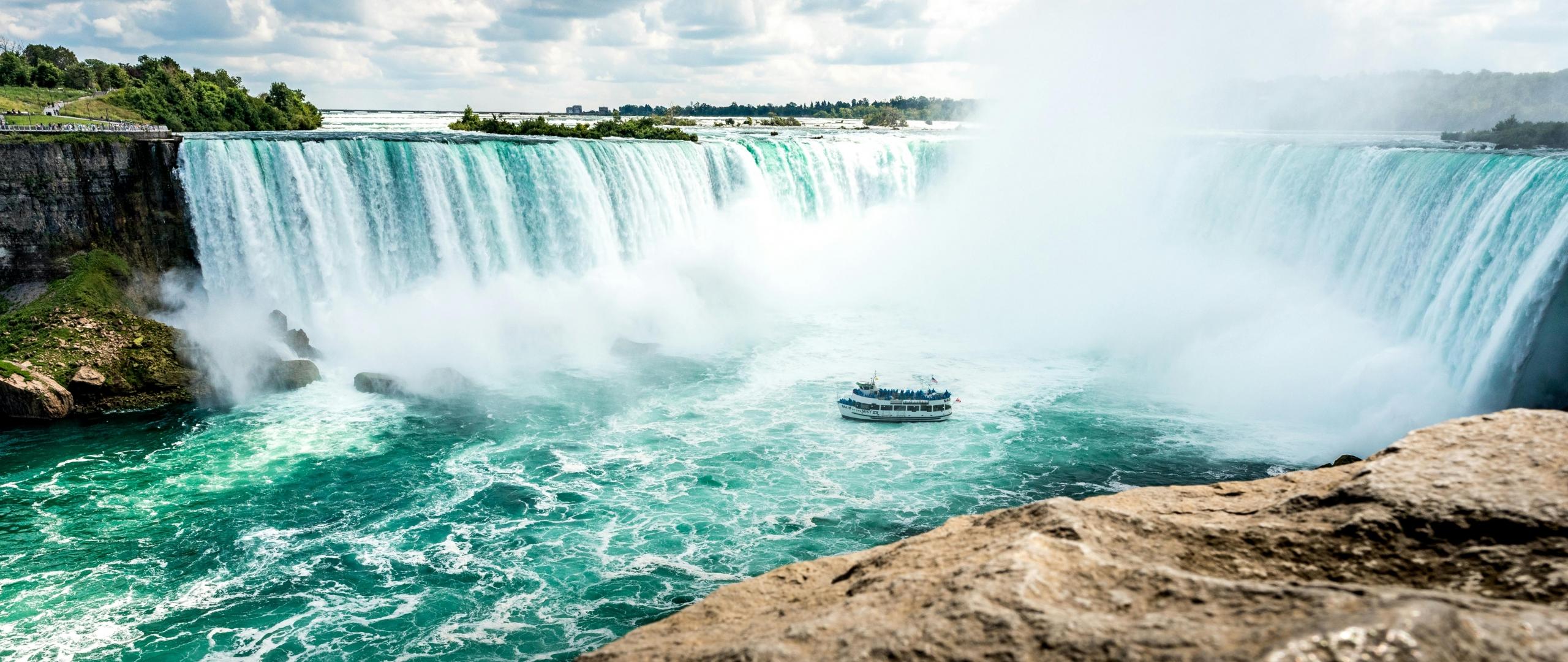 A stunning view of Niagara Falls with the Horseshoe Falls in the background, and a tour boat approaching the base of the falls amidst the mist. The scene includes rocky cliffs and greenery under a partly cloudy sky.
