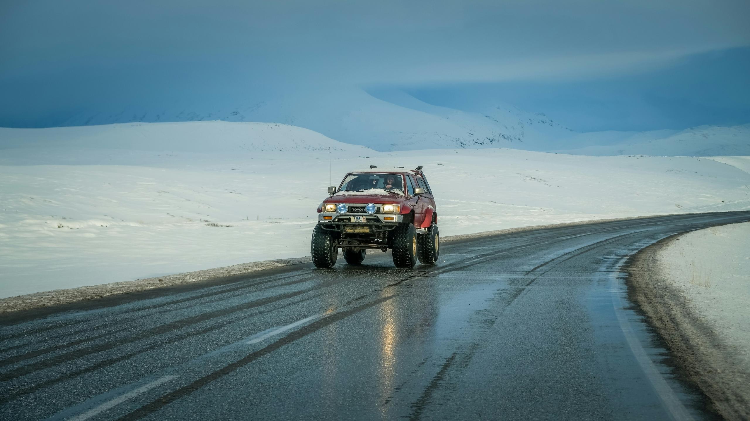 A vehicle drives on an icy road. The surrounding landscape is blanketed in snow, with gentle hills and distant mountains under a cloudy sky, highlighting the remote and harsh beauty of the environment.