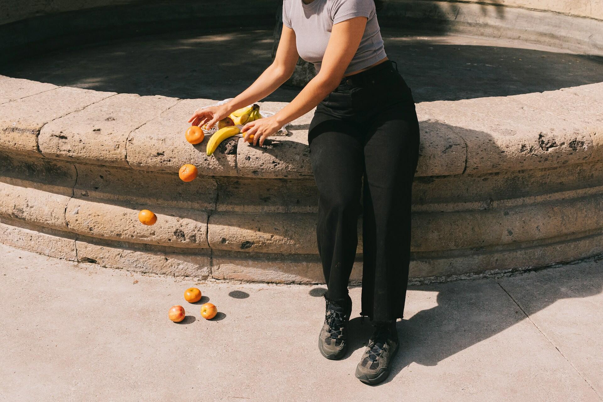 A person wearing a gray top and black pants holds bananas and drops oranges from a stone ledge, with fruit scattered on the ground.