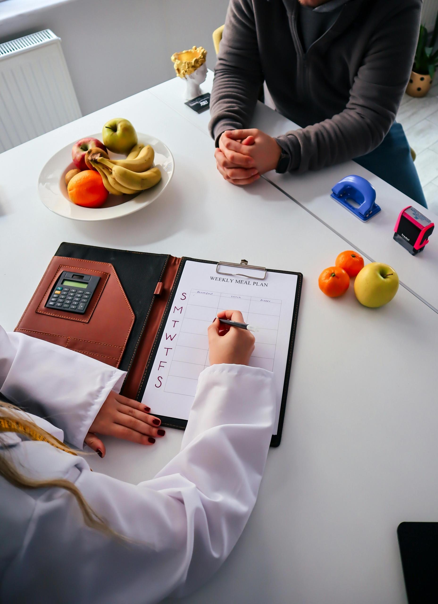 A nutritionist in a white coat writing a weekly meal plan for a client during a consultation. Fresh fruits like bananas, apples, and oranges are placed on the table, along with a calculator and office supplies.